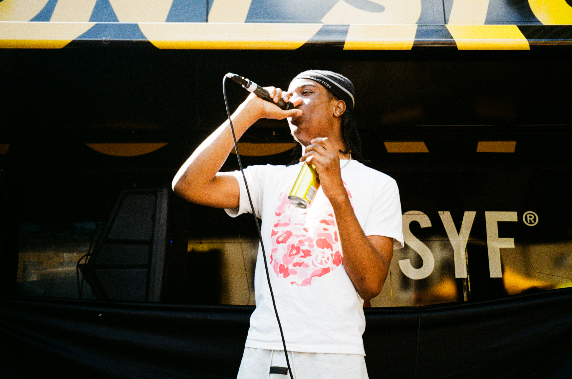 Black woman in white top and headband singing into microphone, yellow and blue striped canopy above, dark storefront behind.