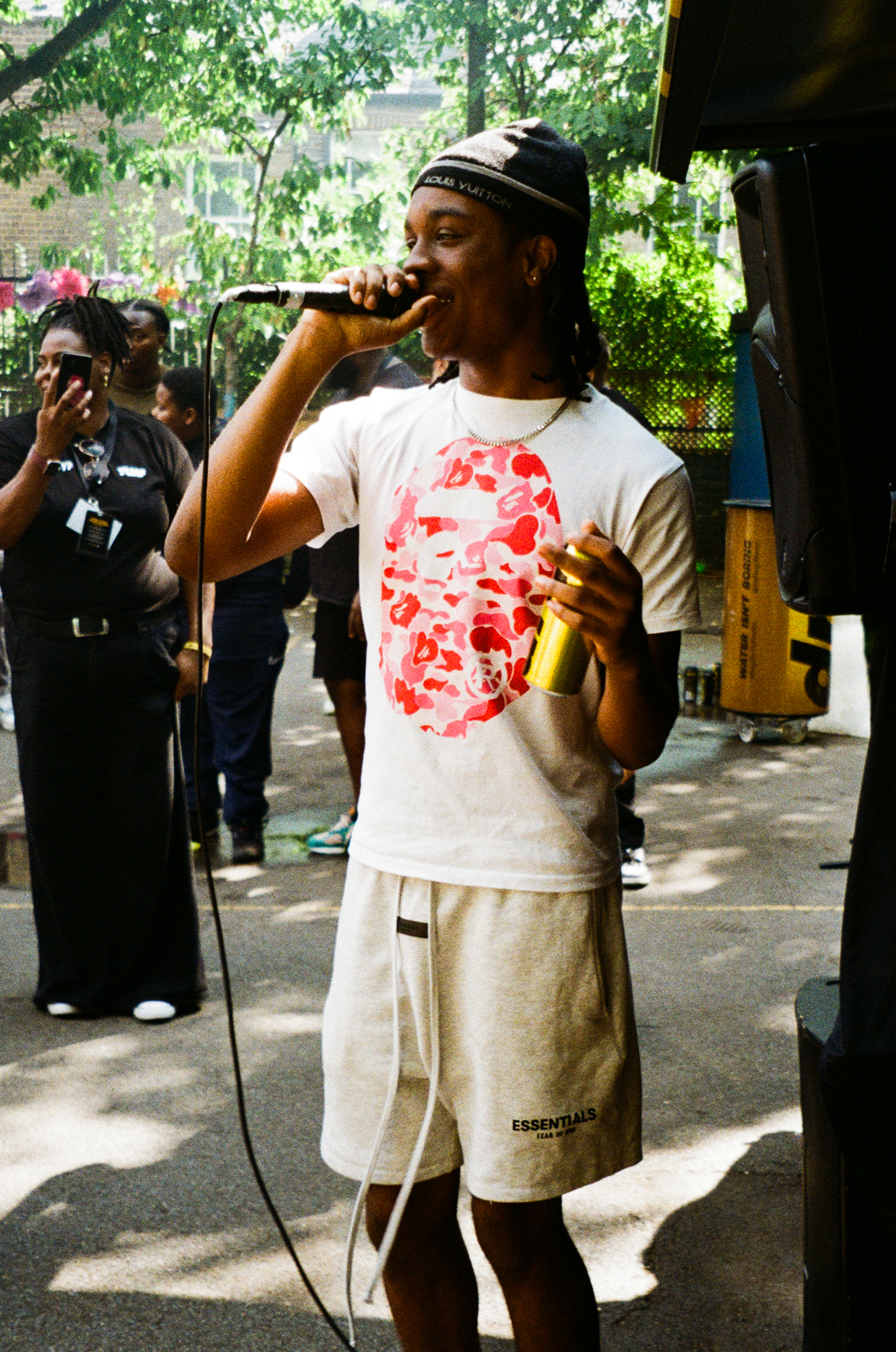 Person with dreadlocks performing with microphone outdoors, wearing white t-shirt with red graphic and beige shorts, crowd behind