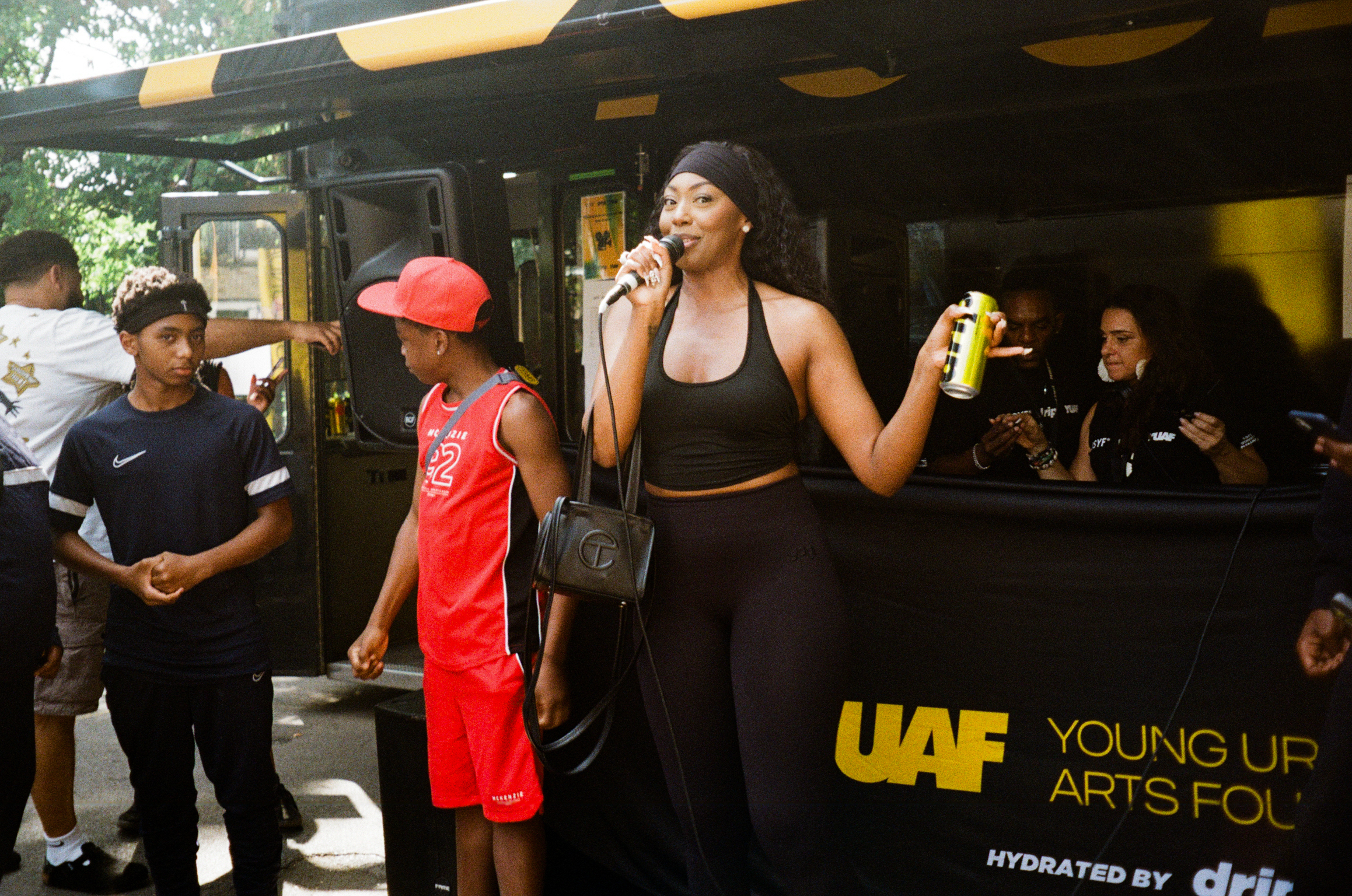 Woman in black top speaking into microphone at outdoor event with children nearby and yellow branded banner visible.