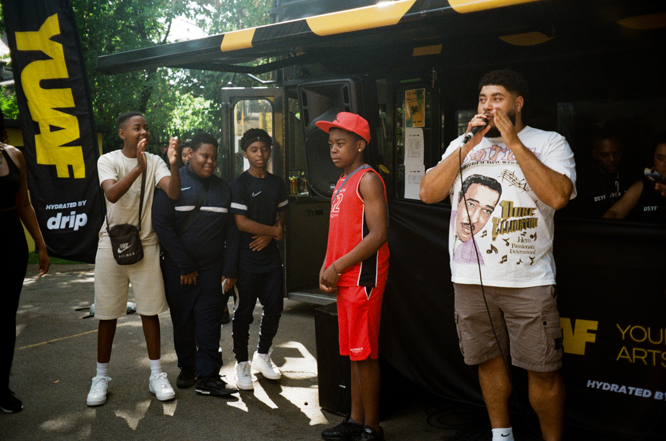 Group of young men performing with microphones at outdoor event under yellow and black striped canopy, with branded backdrop visible.