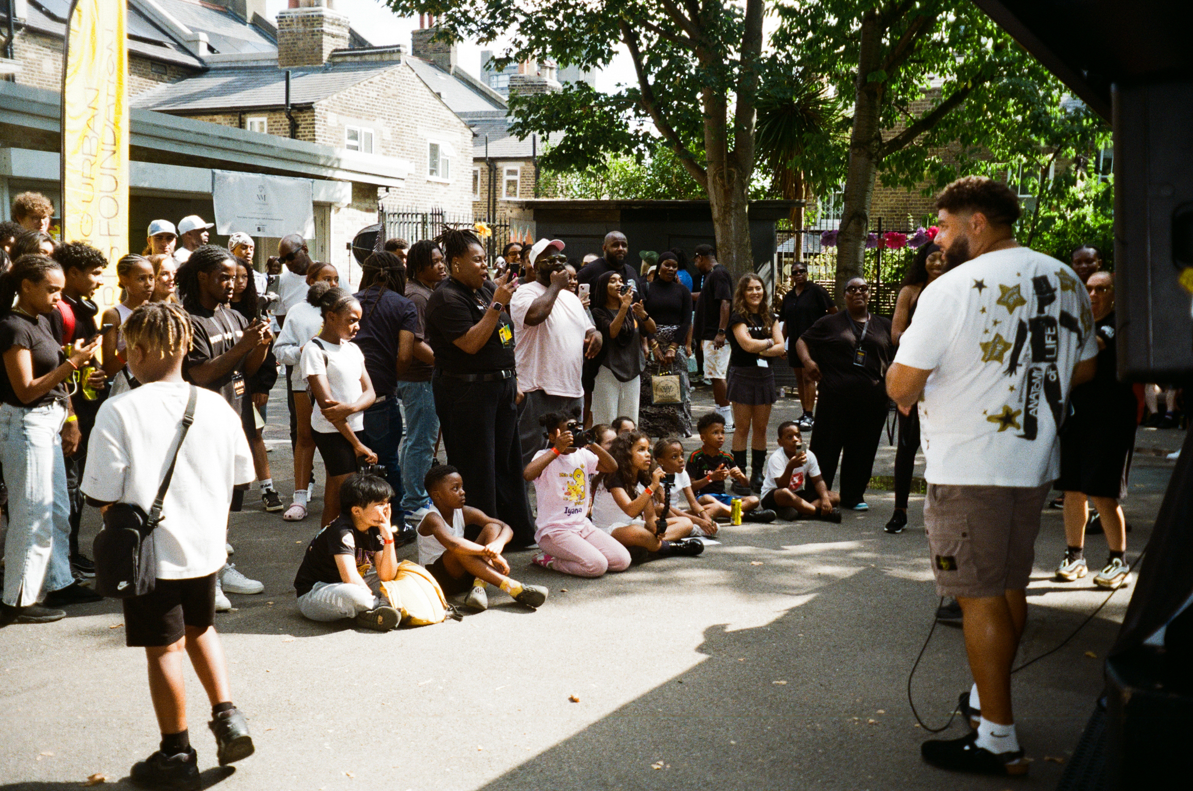 Large crowd gathered in urban plaza with trees and buildings. People standing and sitting on concrete ground in casual clothing.