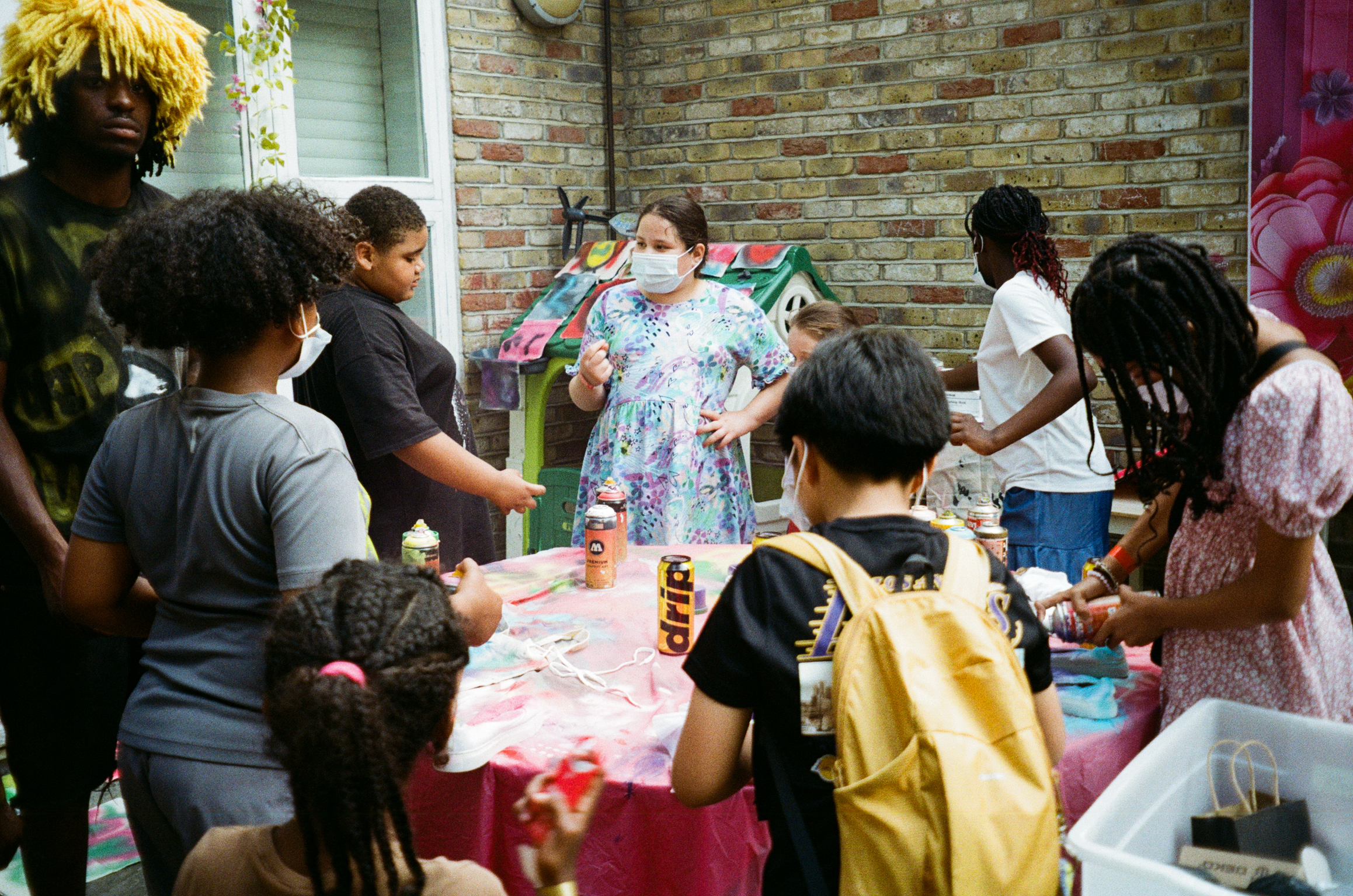 Group of children and adults gathered around tables doing arts and crafts activities indoors against exposed brick wall.