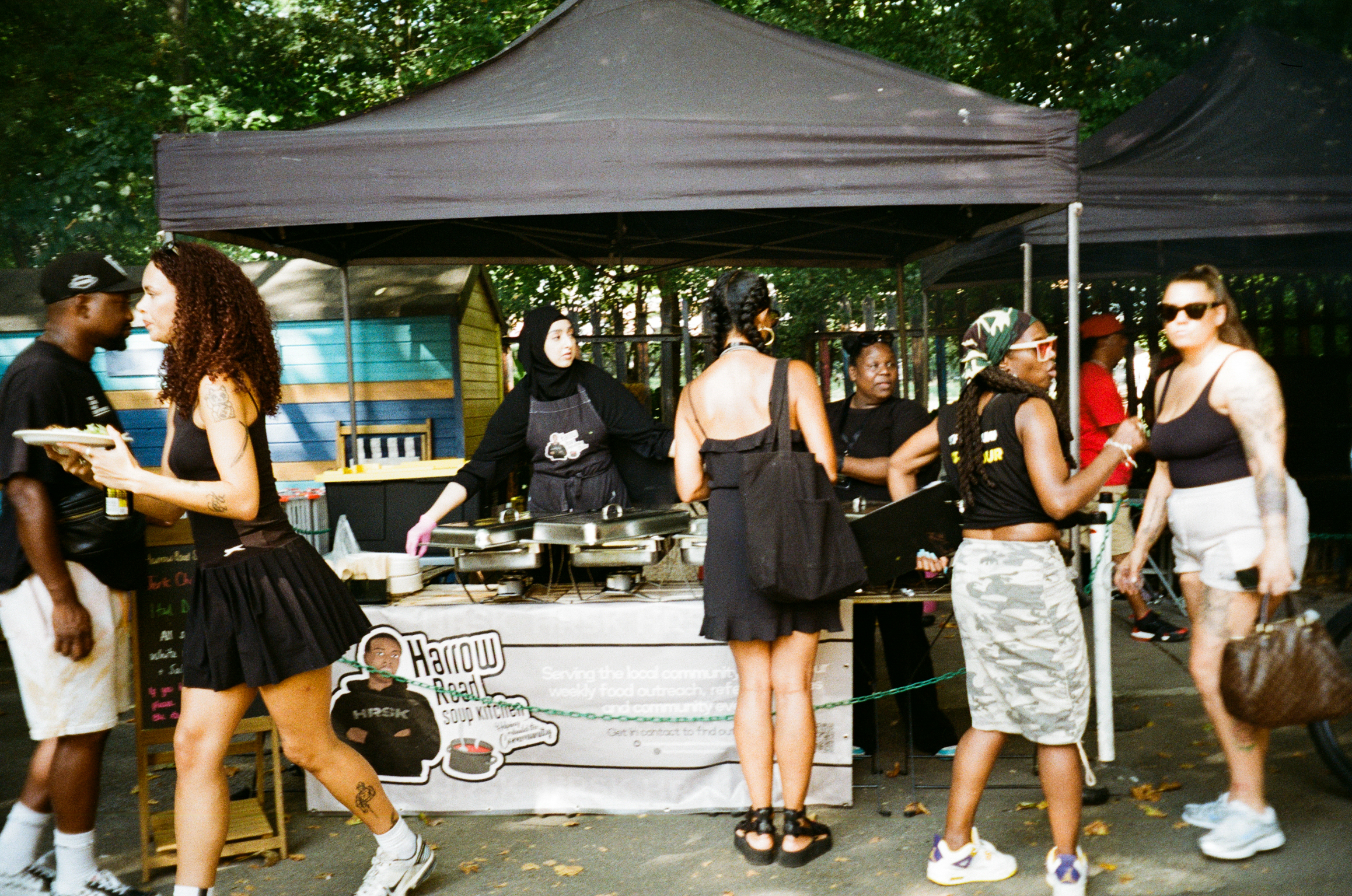 People queuing at outdoor food stall under grey gazebo, staff in black aprons serving from counter with white signage.