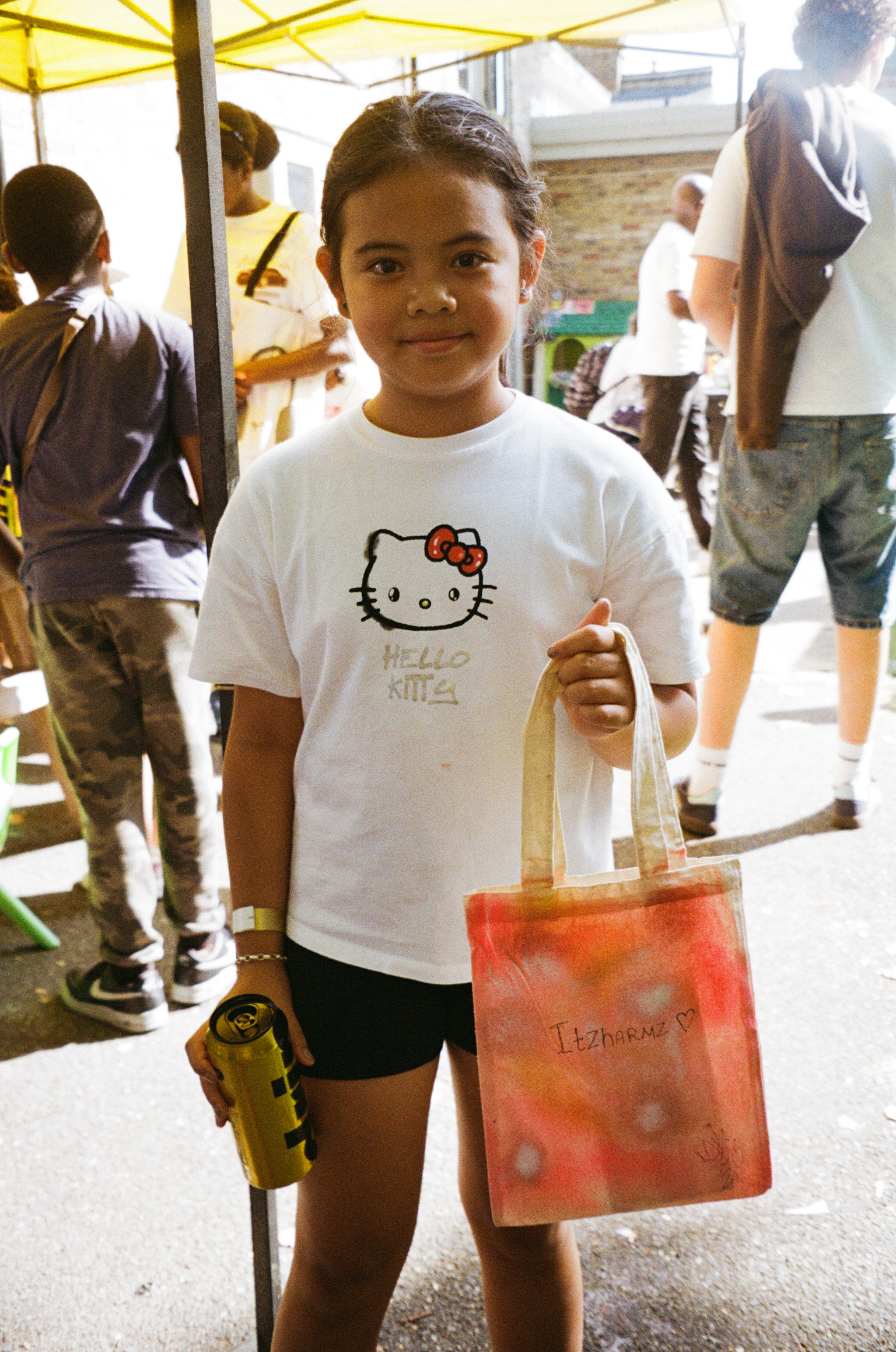 Girl in Hello Kitty t-shirt holds orange shopping bag at outdoor market with yellow canopy and other shoppers in background.