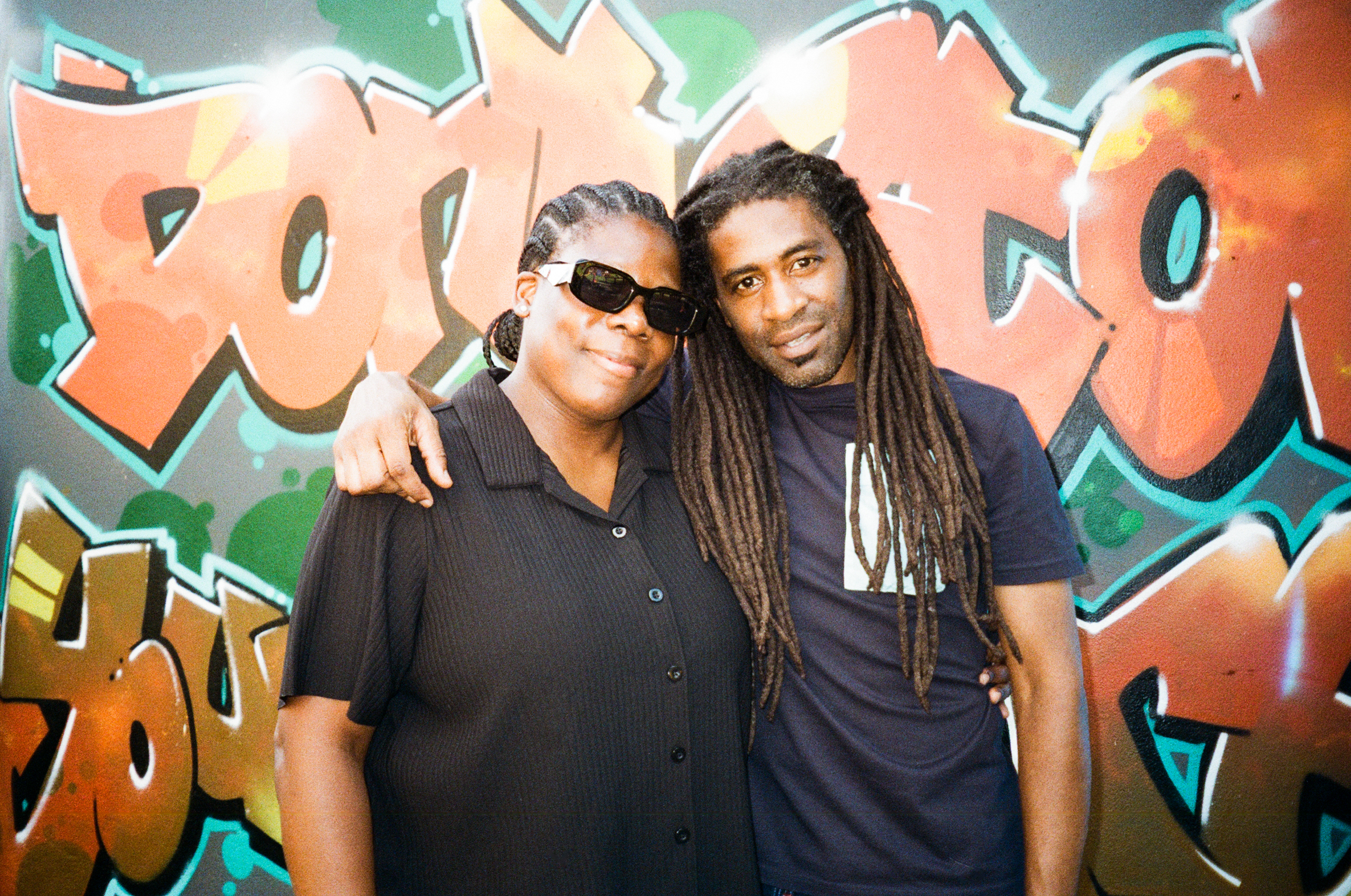 Two men pose together against colourful graffiti wall with orange, green, and teal lettering. One wears sunglasses and black shirt, other has long dreadlocks and dark top.