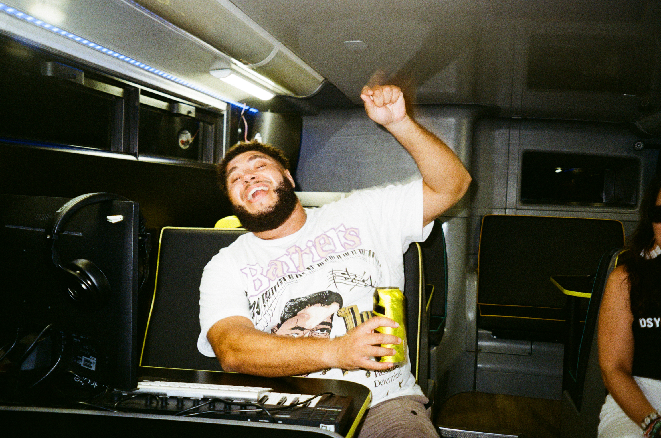 Man with raised fist celebrating in vehicle interior, wearing white graphic t-shirt, holding yellow drink, equipment visible.