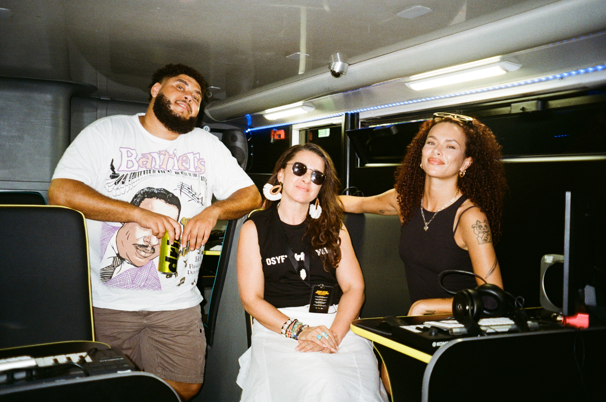 Three people in recording studio: bearded man in white t-shirt, woman in sunglasses and black top seated, blonde woman standing right.