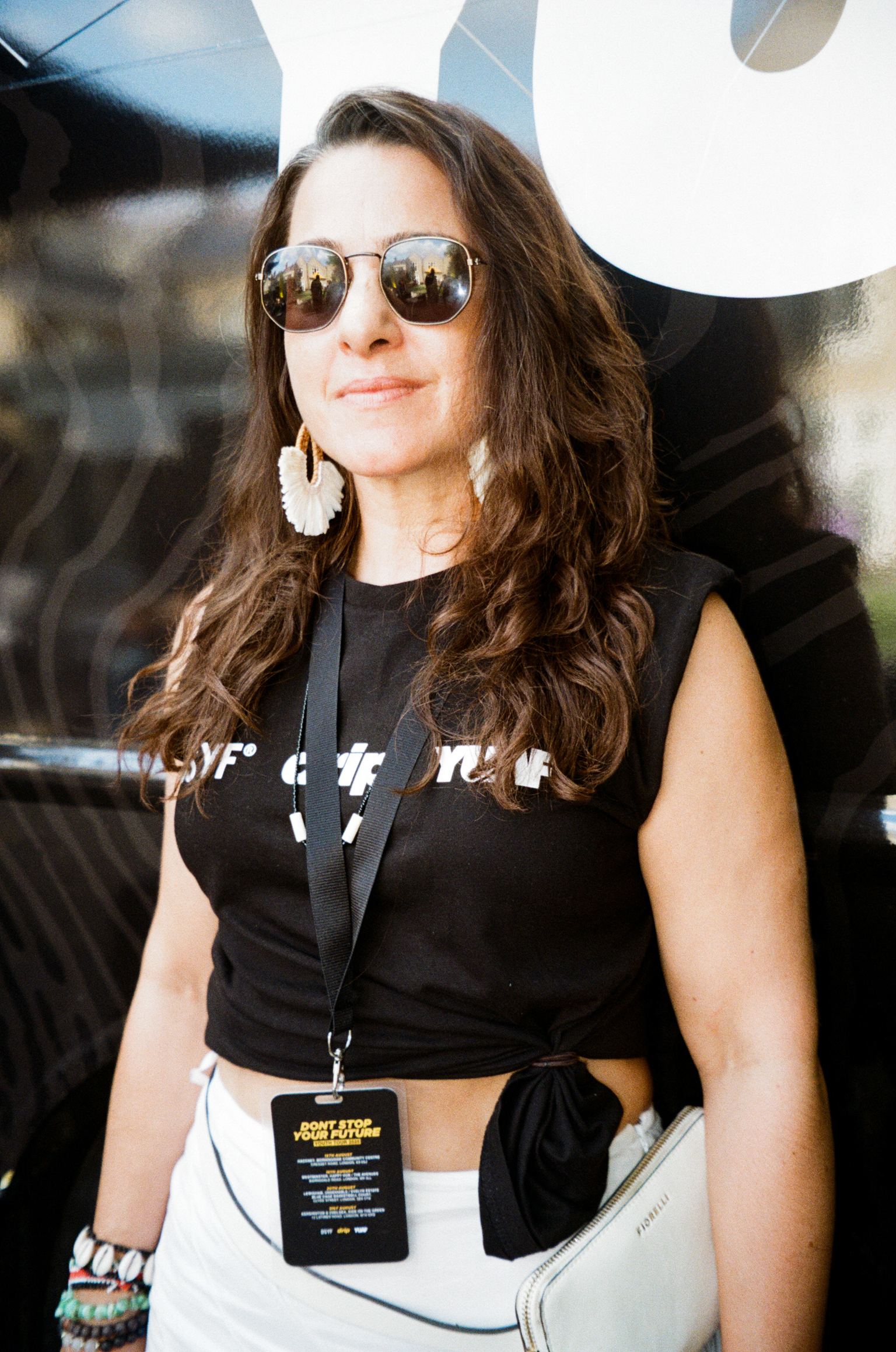 Woman with long brown hair wearing sunglasses, black sleeveless top, white tassel earrings, and lanyard with badge at outdoor event.