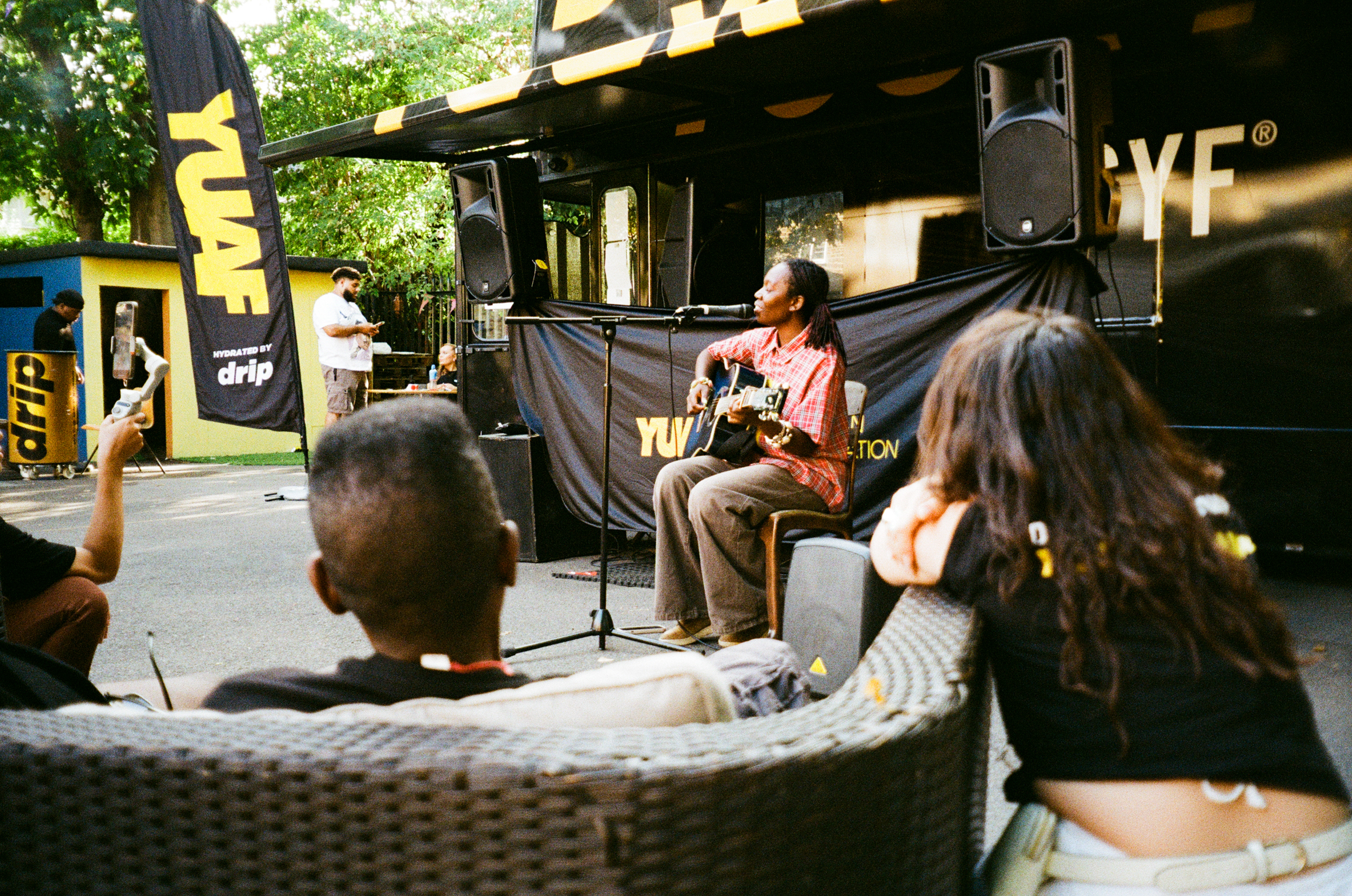 Man in orange shirt playing acoustic guitar on outdoor stage with seated audience on wicker chairs in sunny courtyard setting.