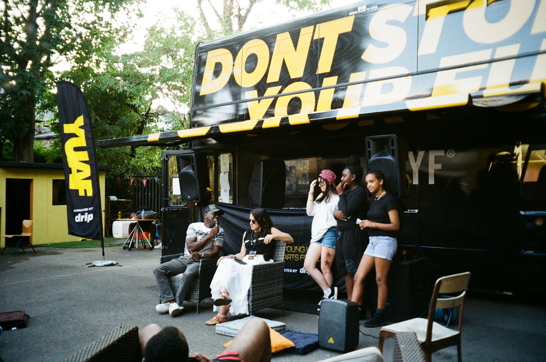 Group of people gathered outside black and yellow building with "DON'T STOP" text on facade, outdoor seating area with chairs visible.