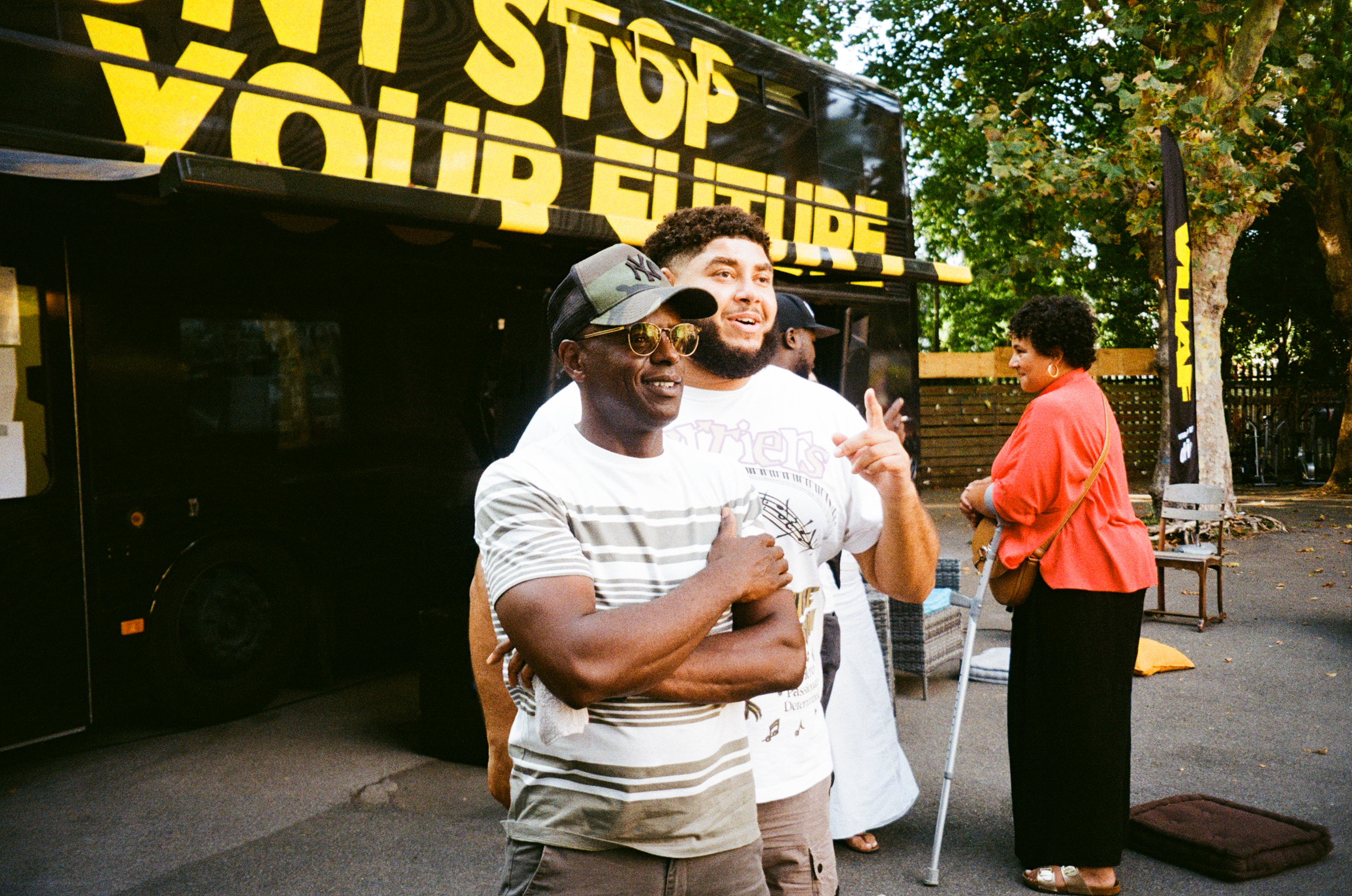Two people embracing in front of black food truck with yellow "STOP YOUR FIRE" text, trees and person in orange shirt visible in background.