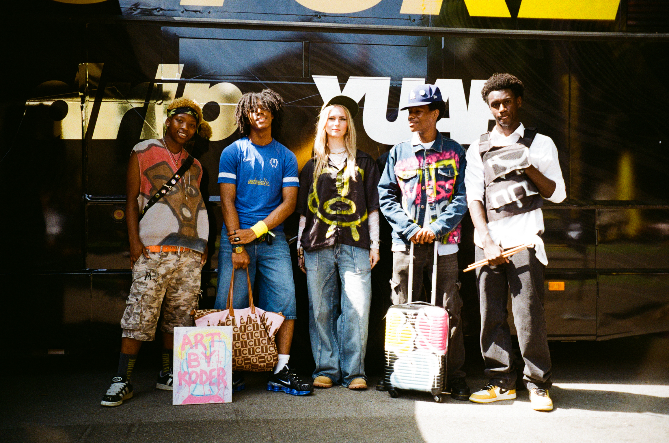Five people standing in front of large blue lorry, holding shopping bags, wearing casual streetwear including jeans and trainers.