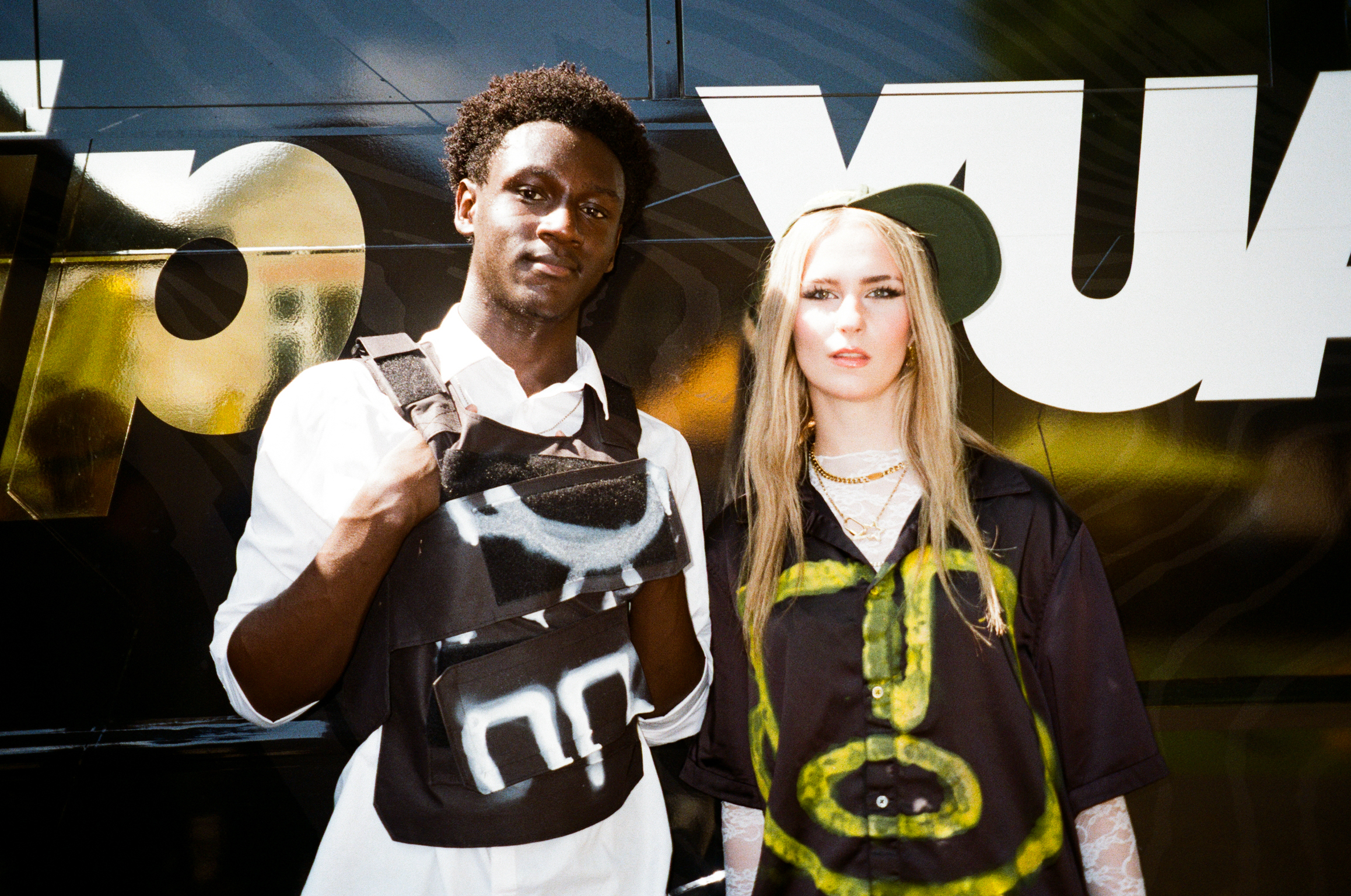 Two young people standing together outdoors; man in white shirt and dark vest, woman in dark top with yellow design and green cap.