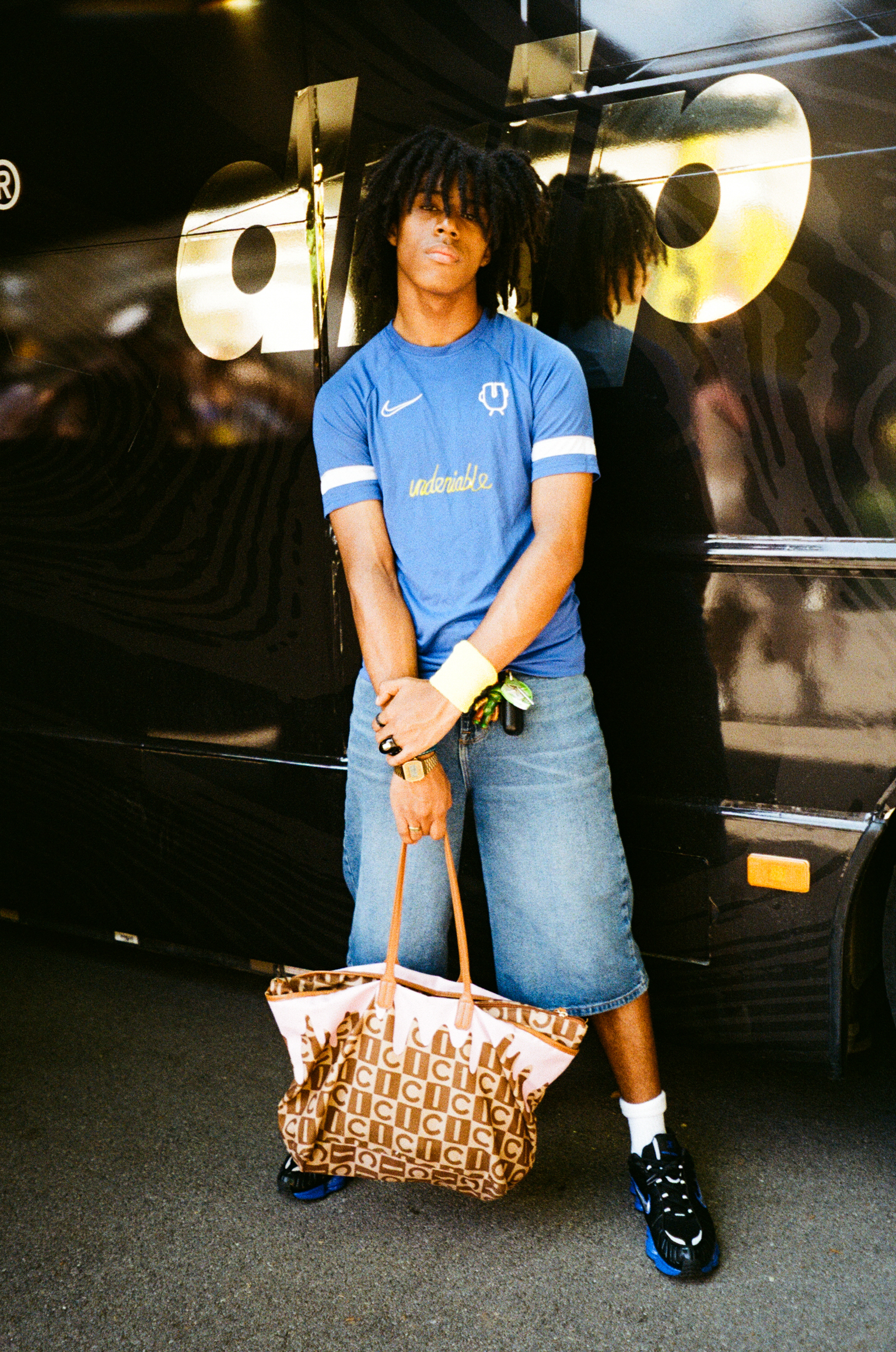 Person with dreadlocks wearing blue football shirt and denim skirt, holding patterned handbag, standing beside black vehicle.
