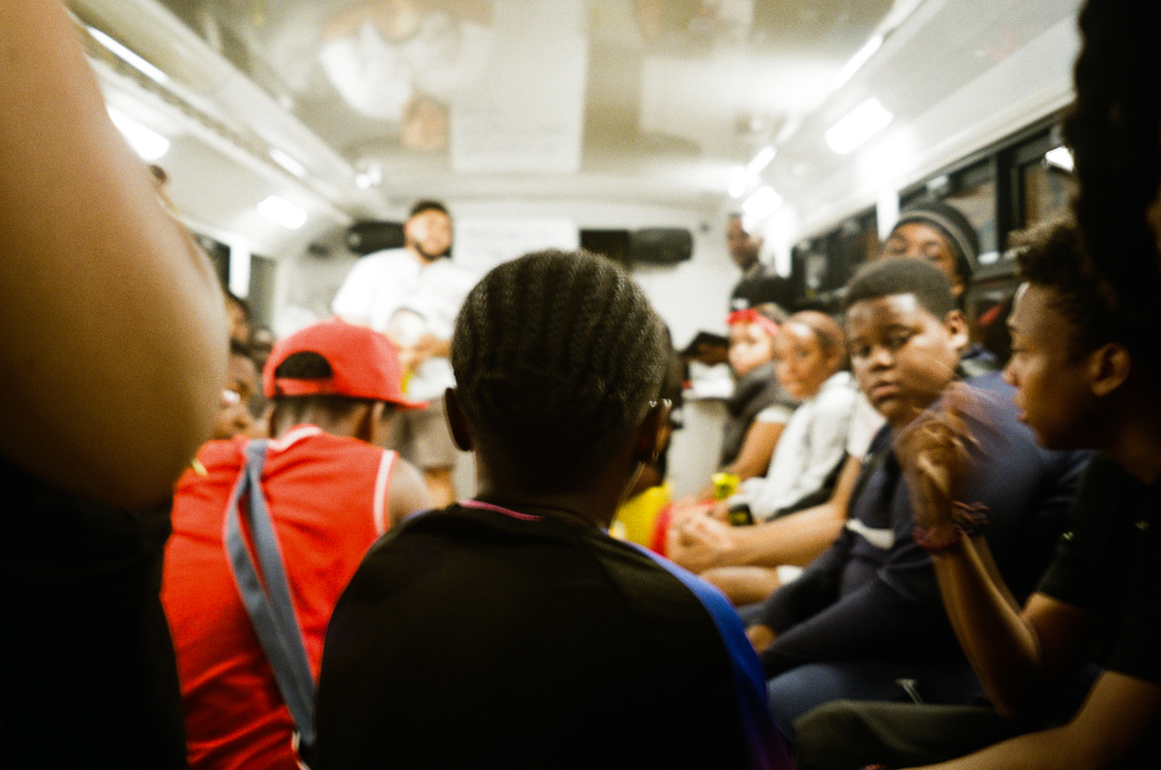 Crowded bus interior with passengers seated and standing, person in bright orange high-visibility jacket and red cap visible amongst commuters.
