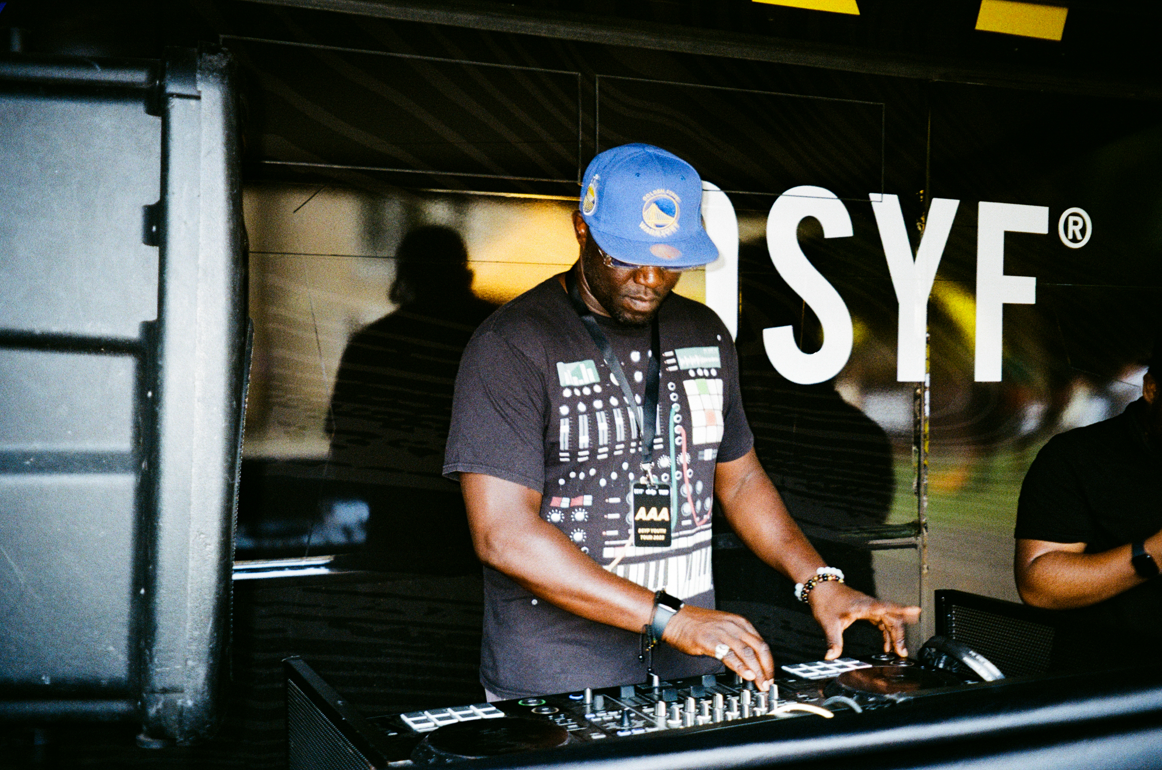 DJ wearing blue cap and black graphic t-shirt operating mixing deck at indoor venue with white "SYF" signage visible behind.
