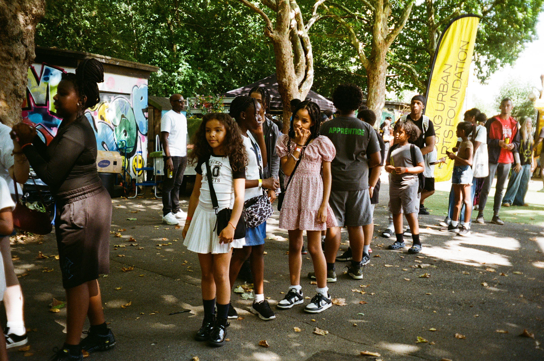Children and adults queuing outdoors under trees, with display boards and yellow banner visible in background on sunny day.