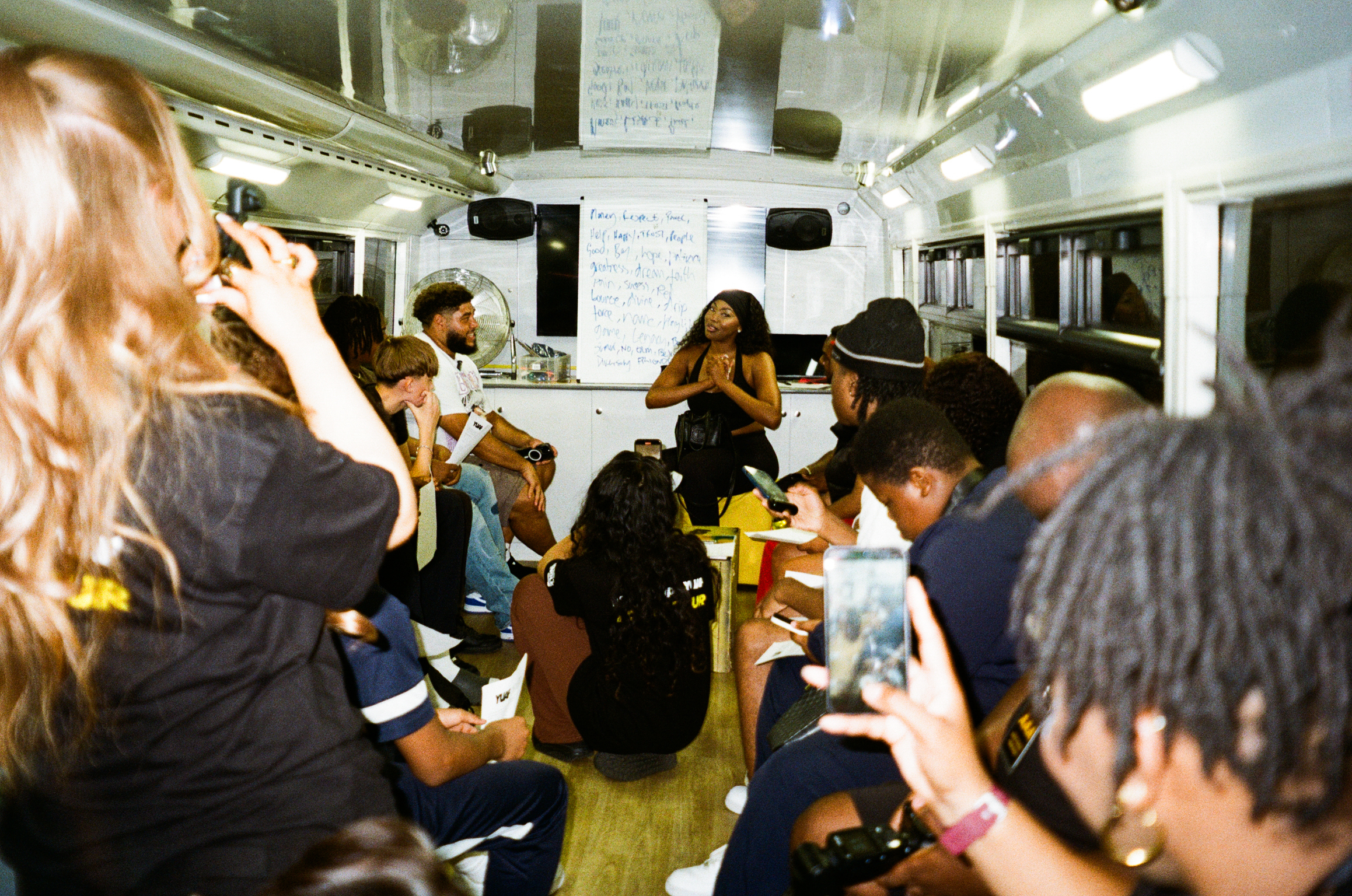 Interior of white mobile unit with multiple people seated and standing, some wearing dark clothing, fluorescent lighting overhead.