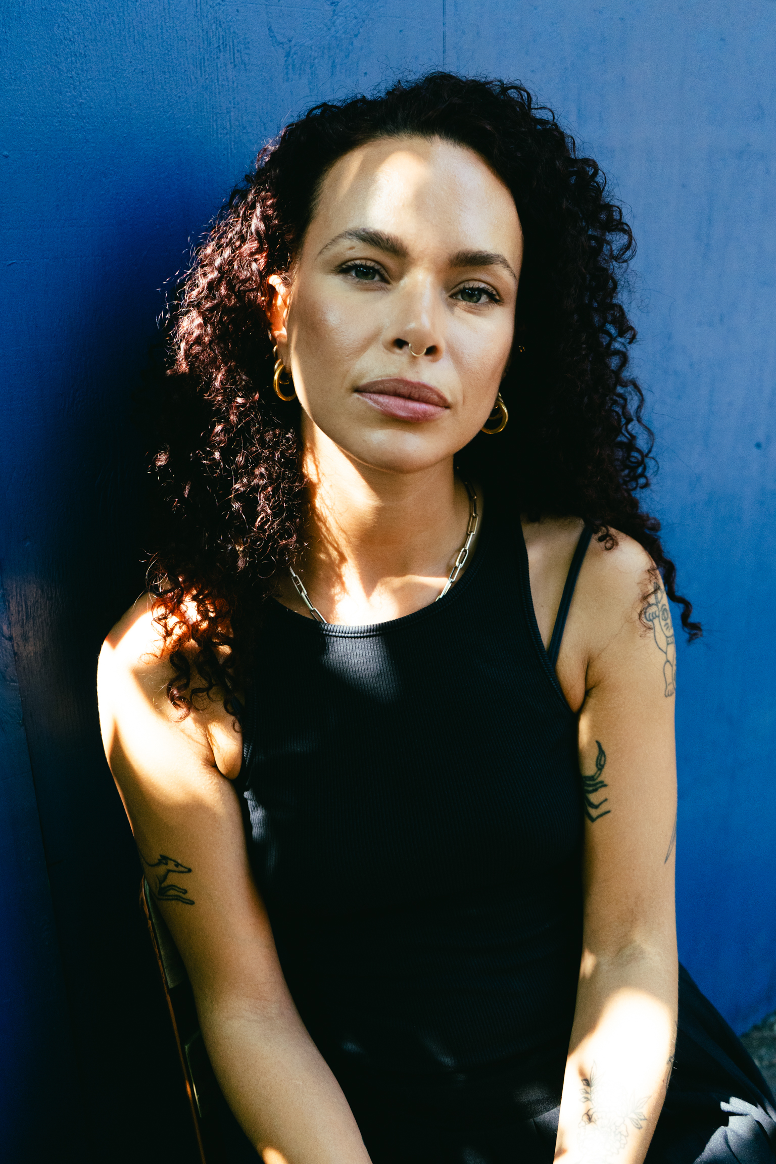 Woman with dark curly hair in black vest top against blue background, sunlight creating shadows across her face and arms with tattoos.