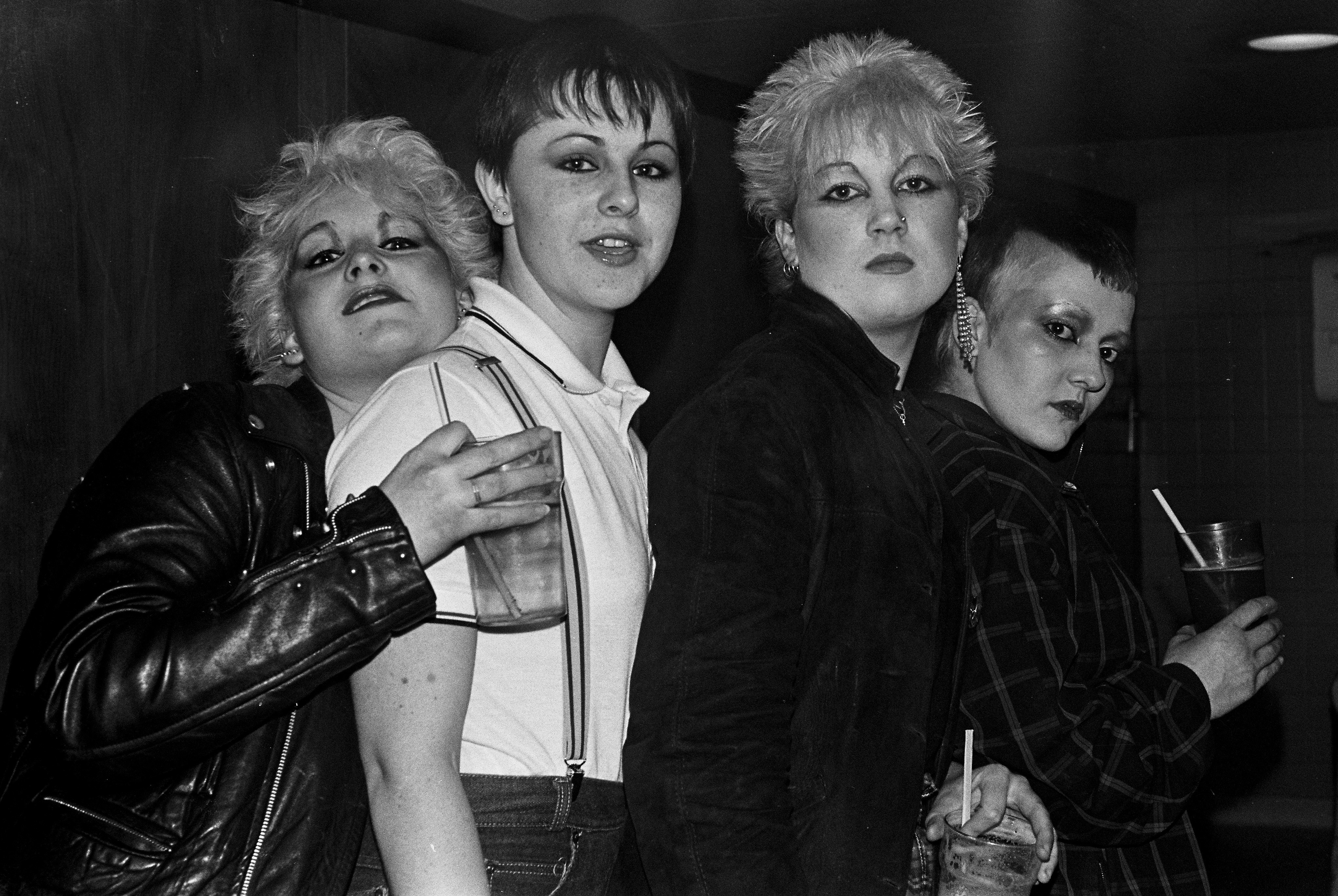 Black and white image of four women with short, spiky blonde hair wearing dark leather jackets, posed closely together.