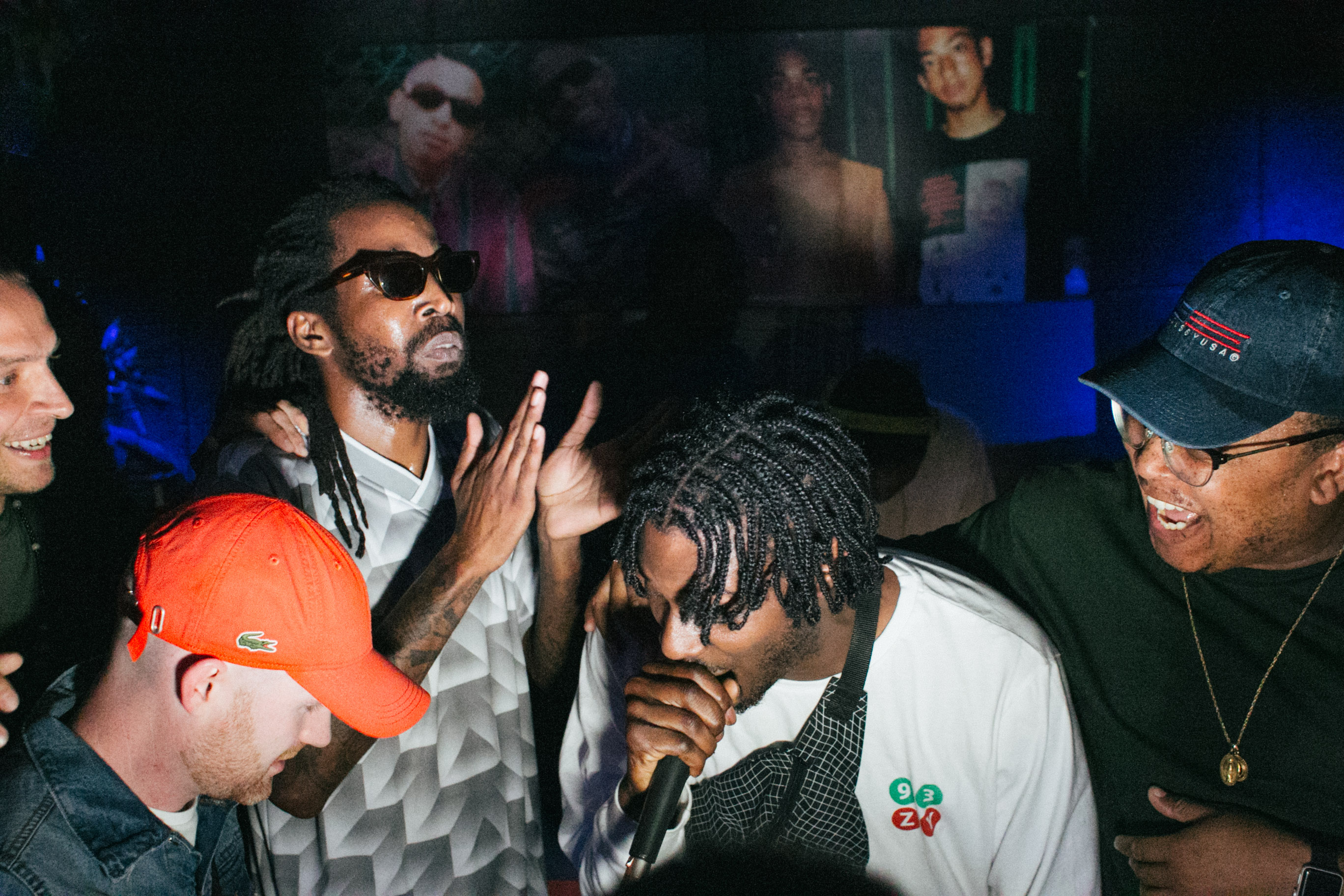 Group of people at nightclub with blue lighting, man in sunglasses and dreadlocks making hand gestures, others wearing caps and casual clothing.