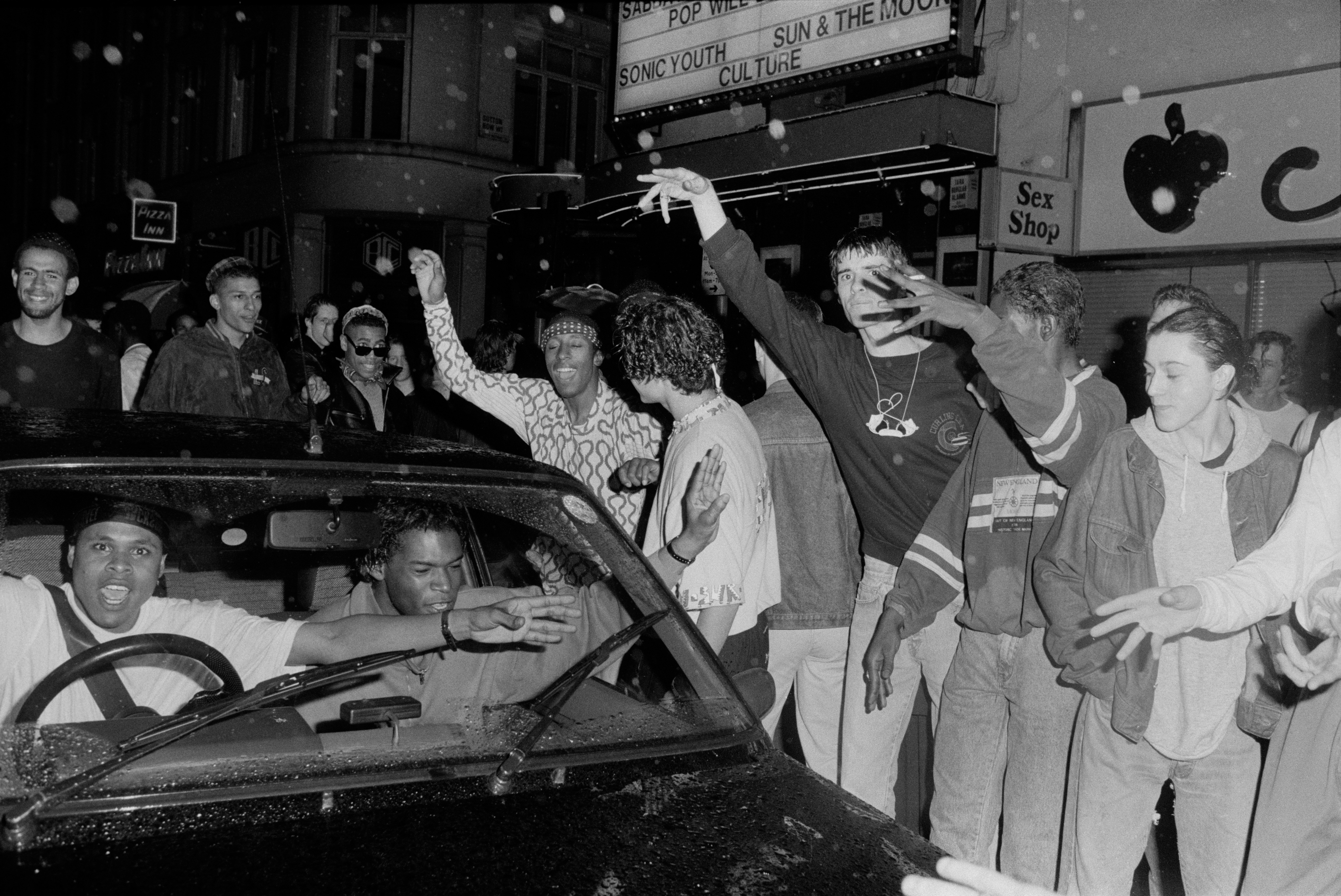 Black and white image showing crowd gathered around convertible car on street at night, with storefronts and signage visible in background.