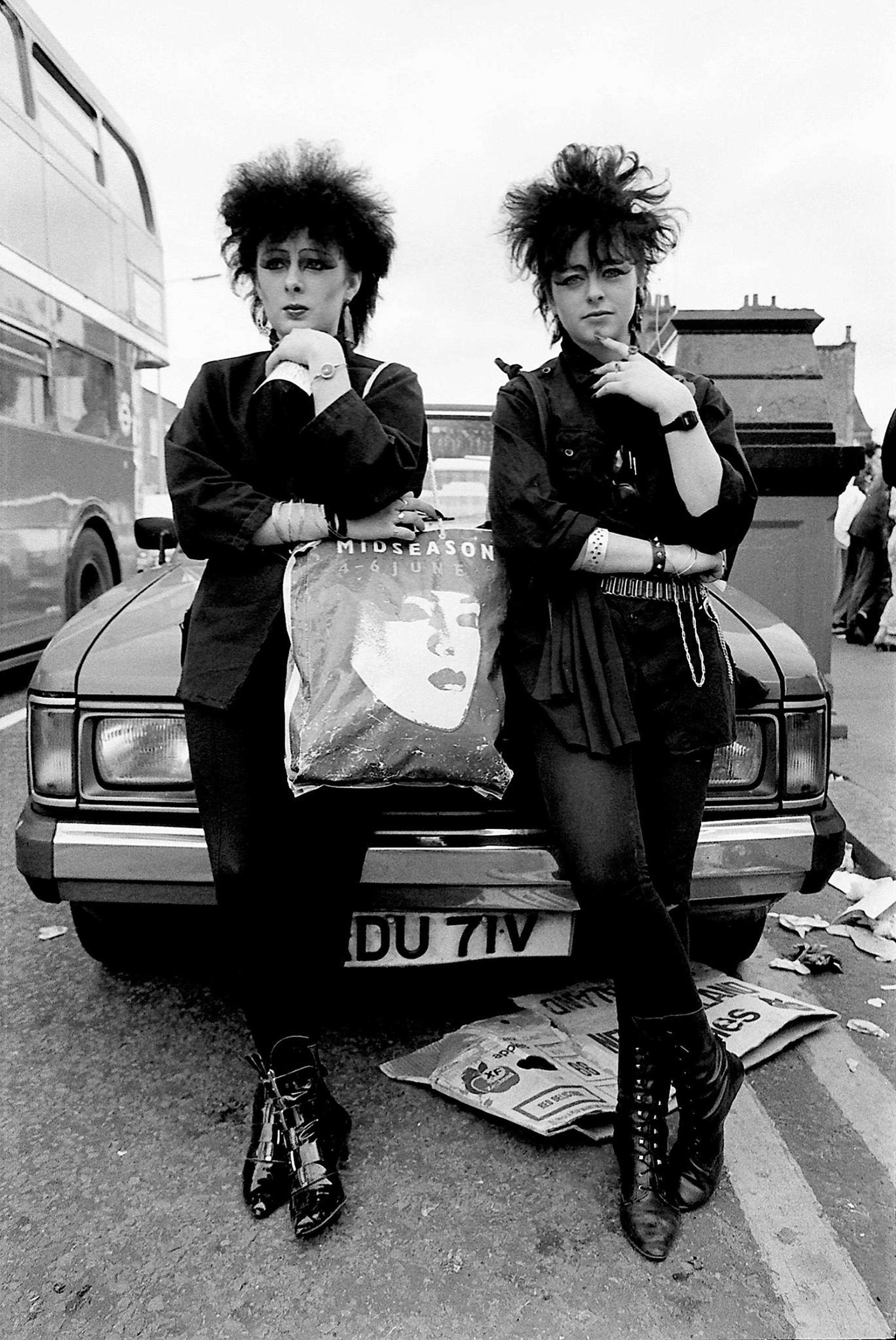 Two people with spiky dark hair in black clothing lean against car bonnet, newspapers scattered on ground, black and white image.