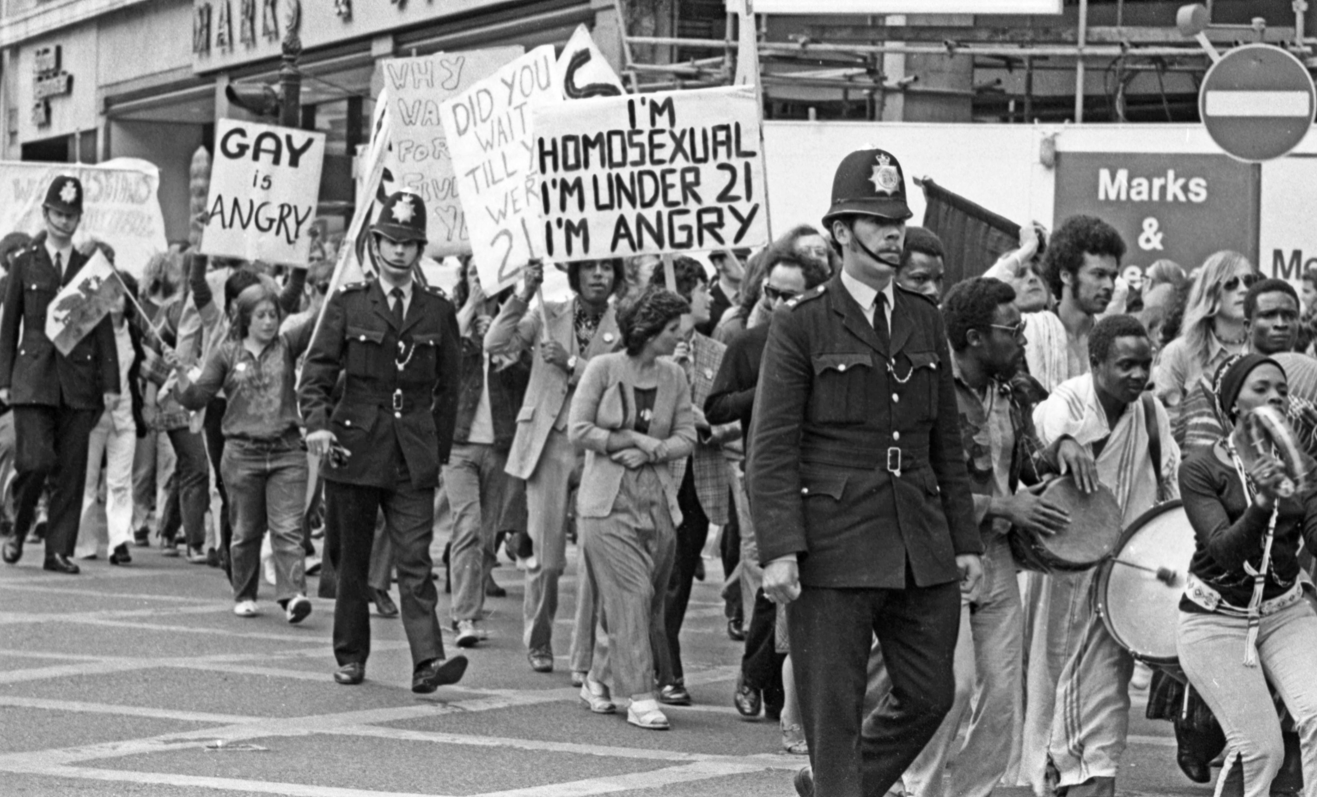 Black and white image of uniformed police officers walking alongside protesters holding signs reading "GAY AND ANGRY" and "HOMOSEXUAL I'M UNDER 21 I'M ANGRY" on city street.