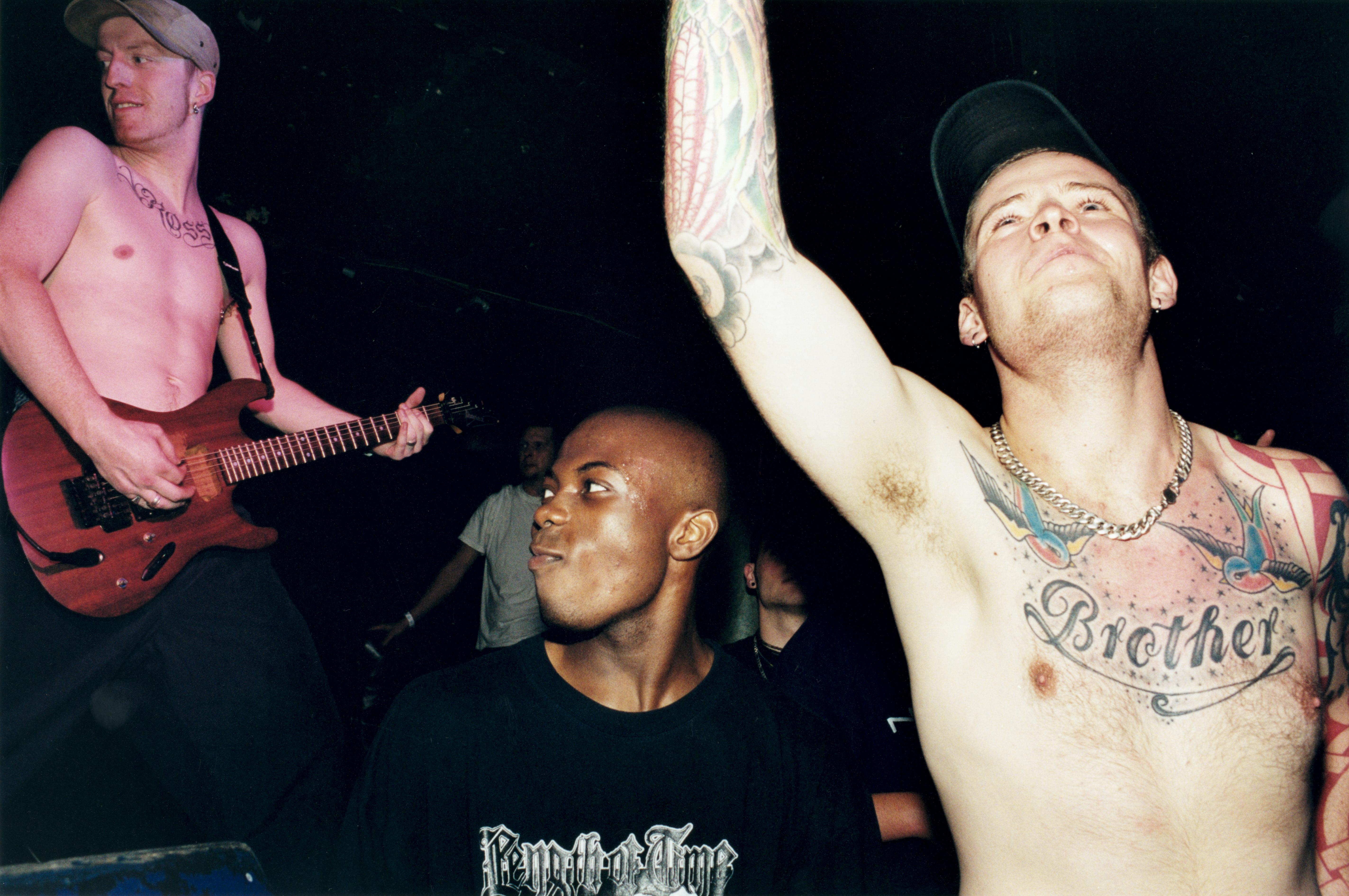 Shirtless tattooed man with arm raised next to Black man in dark shirt, guitarist visible in background at concert venue.