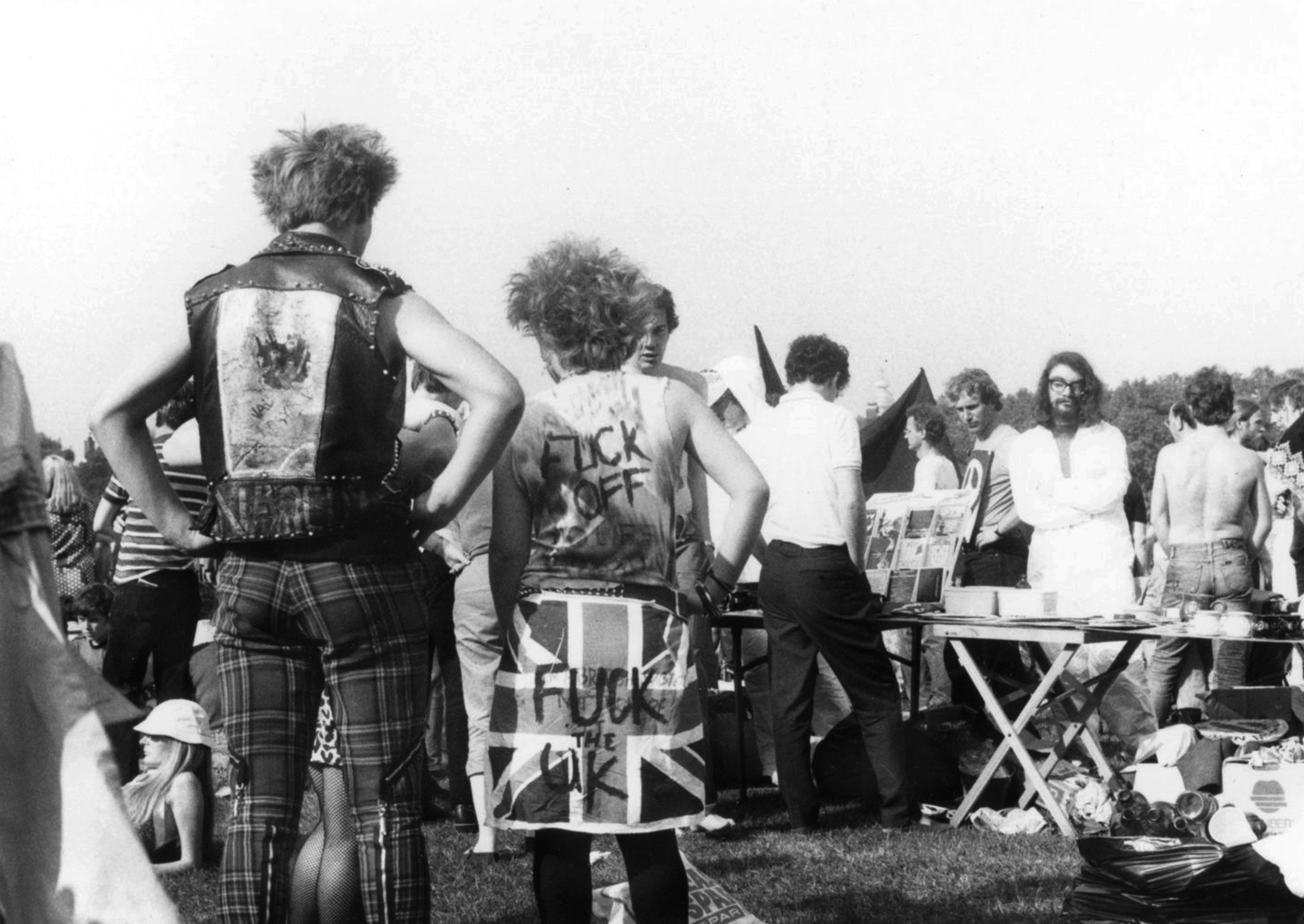 Black and white image showing punks with mohawks and studded leather jackets facing away, crowd gathered around outdoor stall or table.