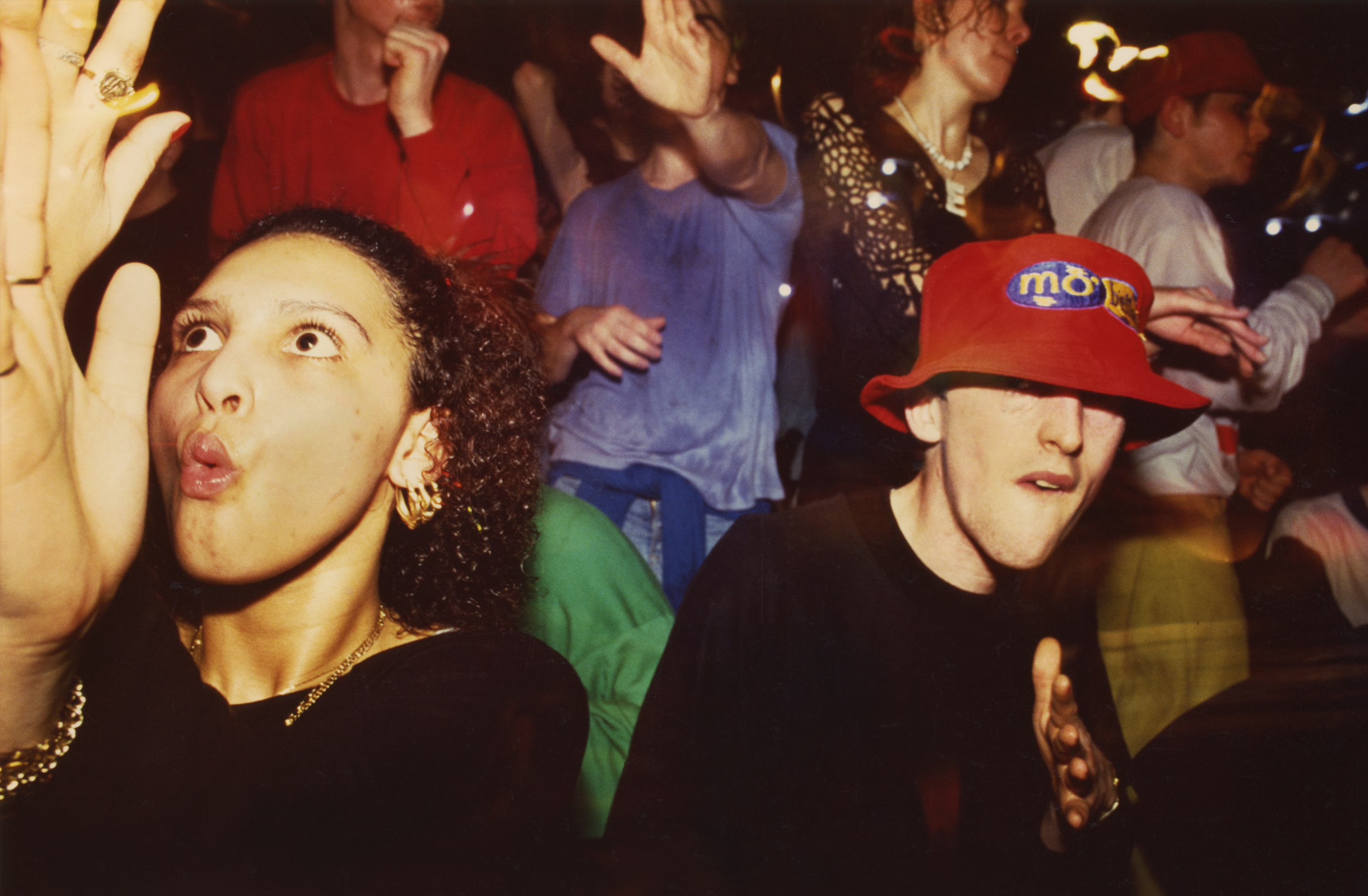 Woman with surprised expression next to person in red cap, surrounded by crowd at nighttime event.