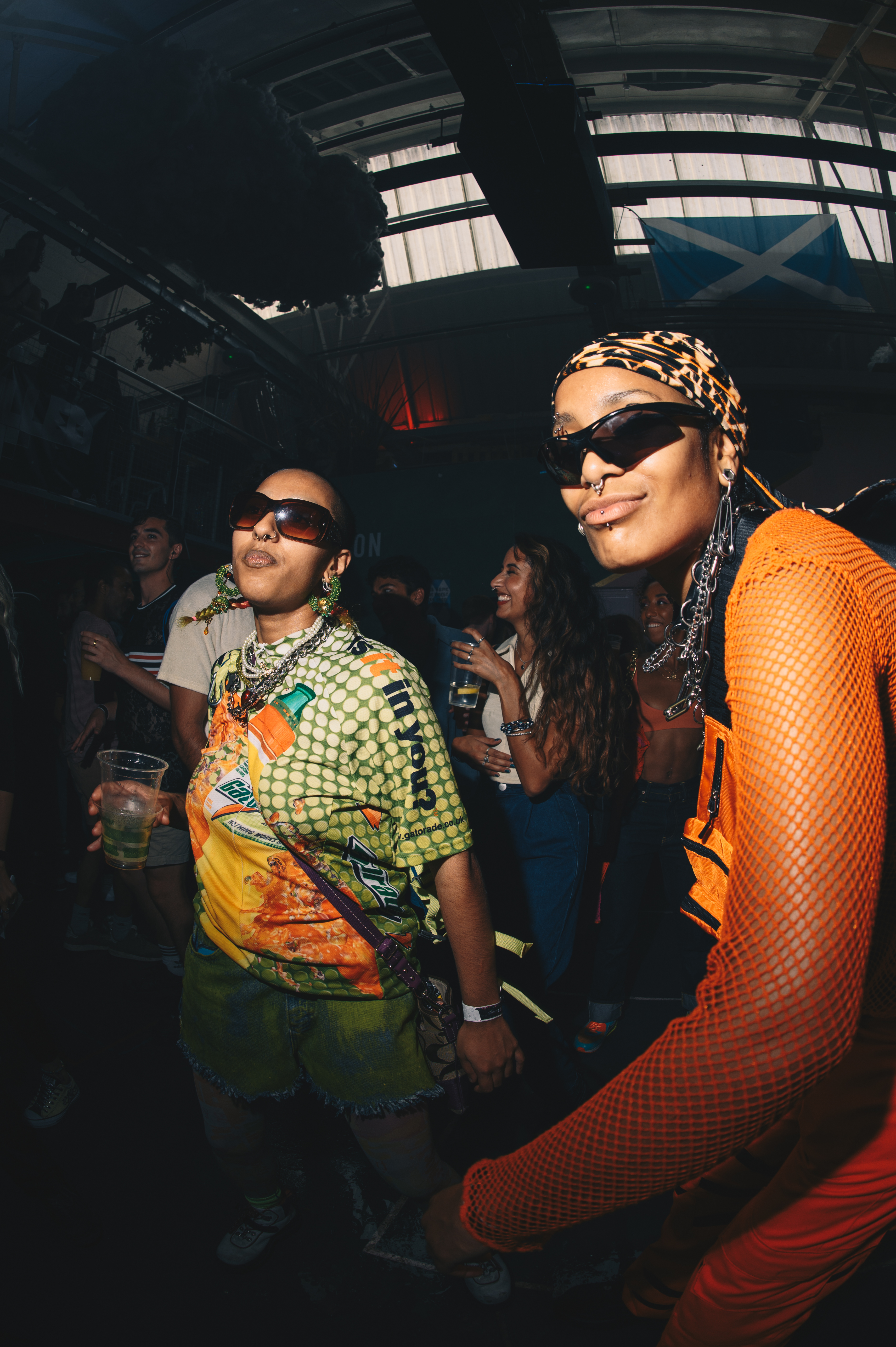 Two people wearing sunglasses at indoor event. Left person in patterned shirt, right person in orange mesh top with headscarf. Dark venue with windows.