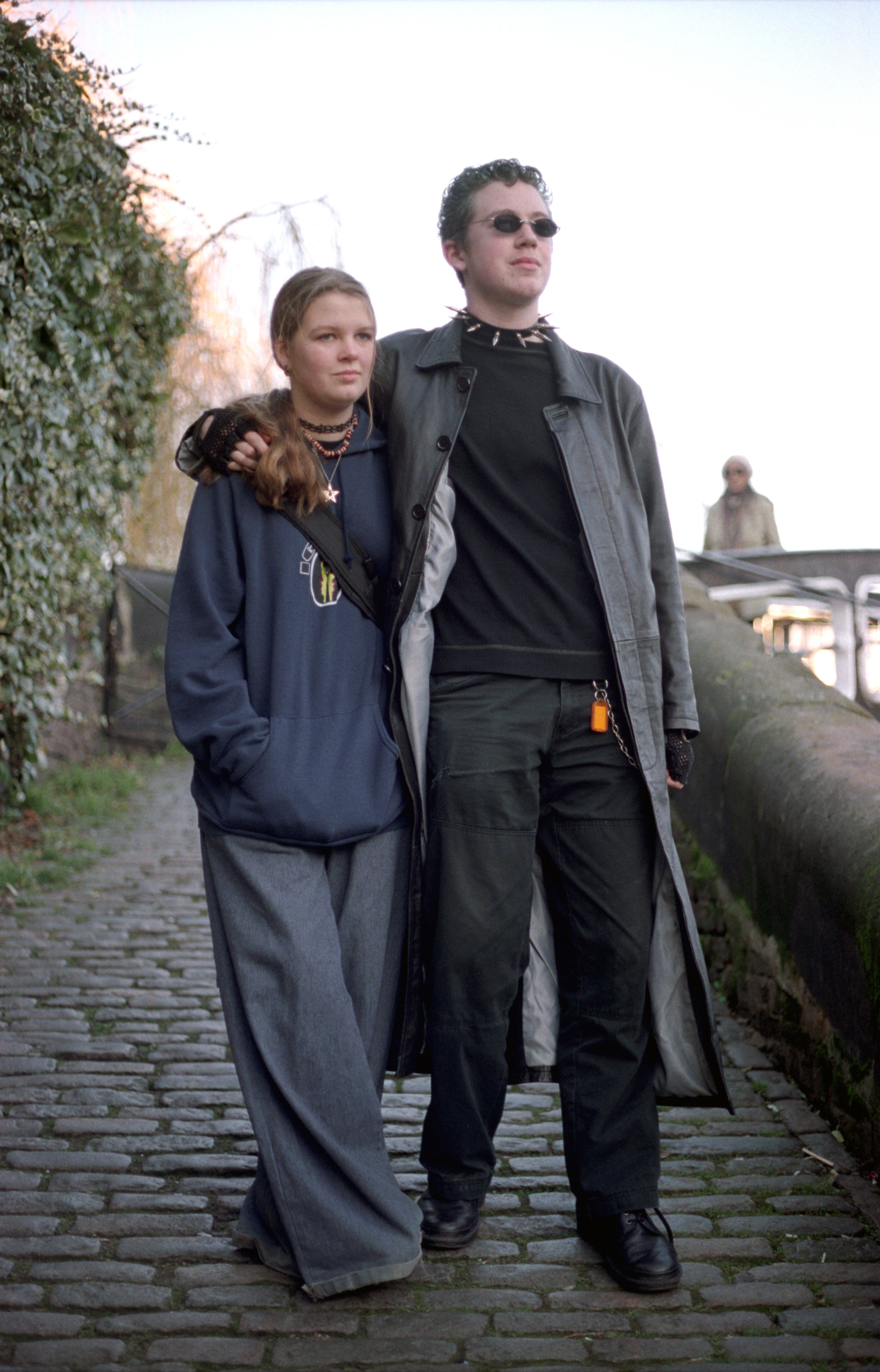 Two people in dark clothing standing on cobblestone street, one wearing sunglasses and long coat, the other in oversized jacket.