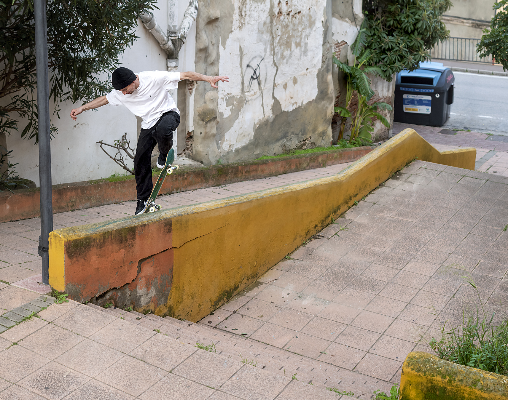 Person skateboarding on yellow concrete ledge in urban street setting with weathered wall and blue postbox in background.