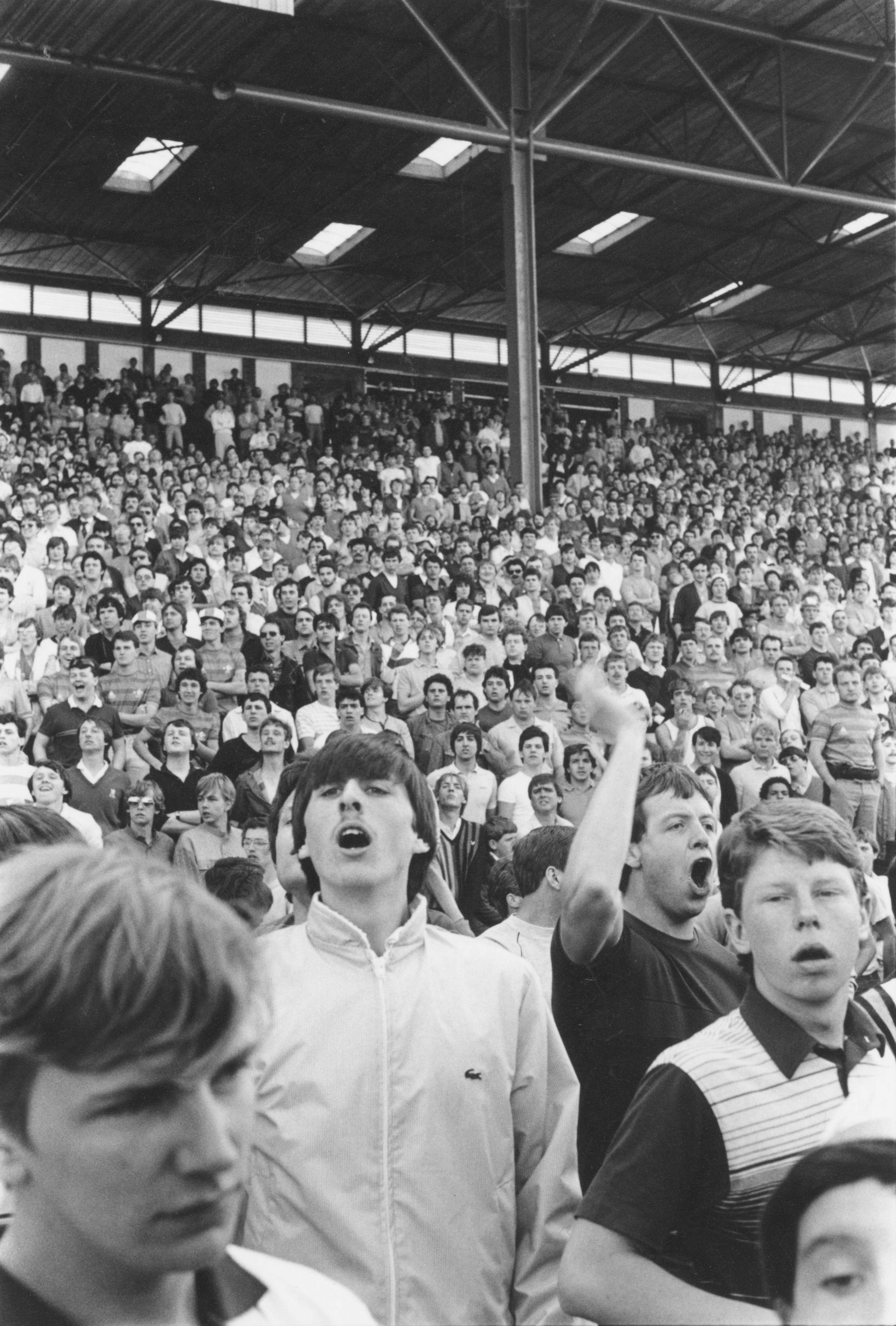Black and white image of crowded stadium terraces with hundreds of spectators, industrial roof structure with skylights visible above.
