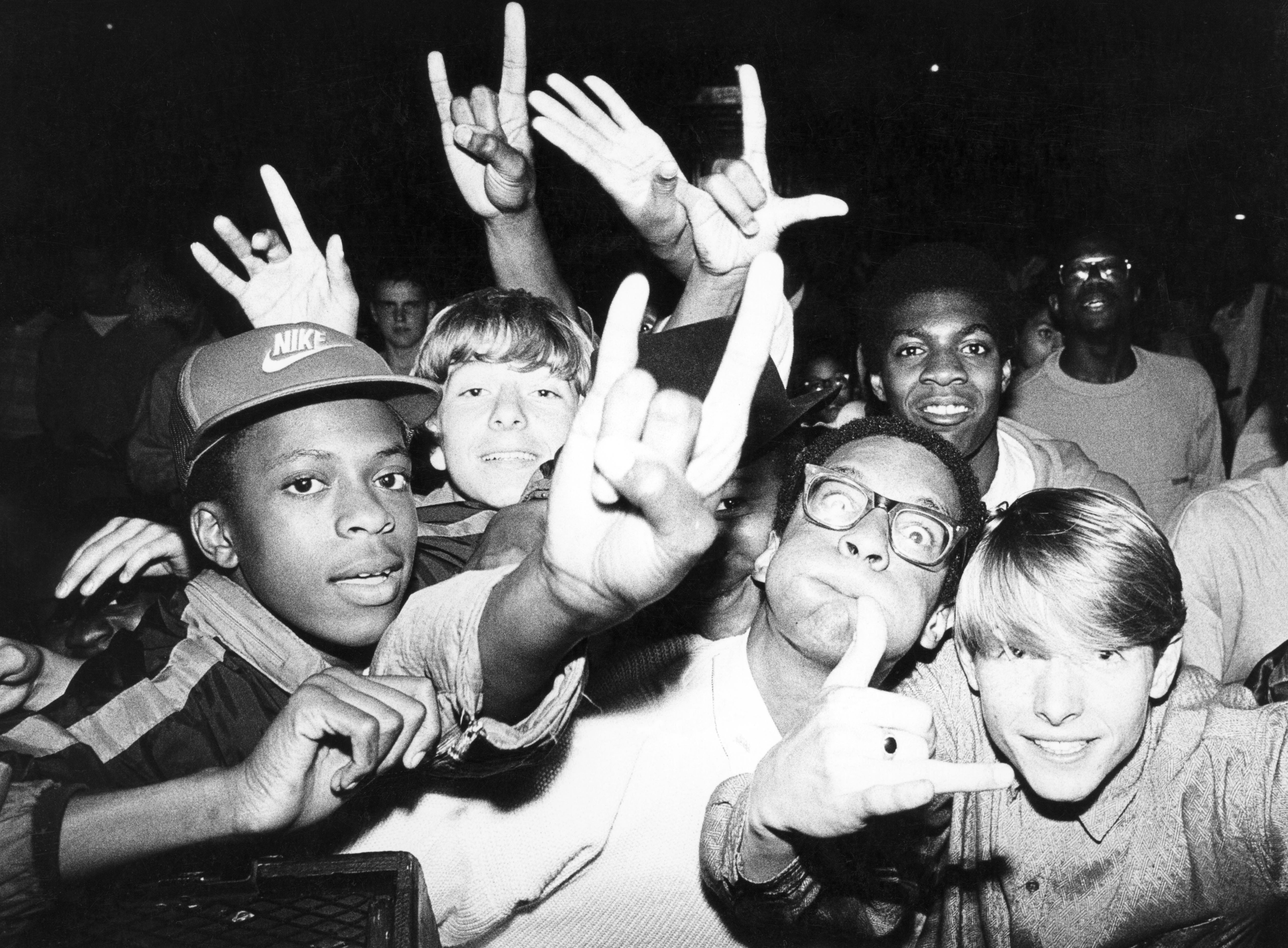 Black and white image of crowd with raised hands making rock horn gestures and peace signs, faces looking upward excitedly.
