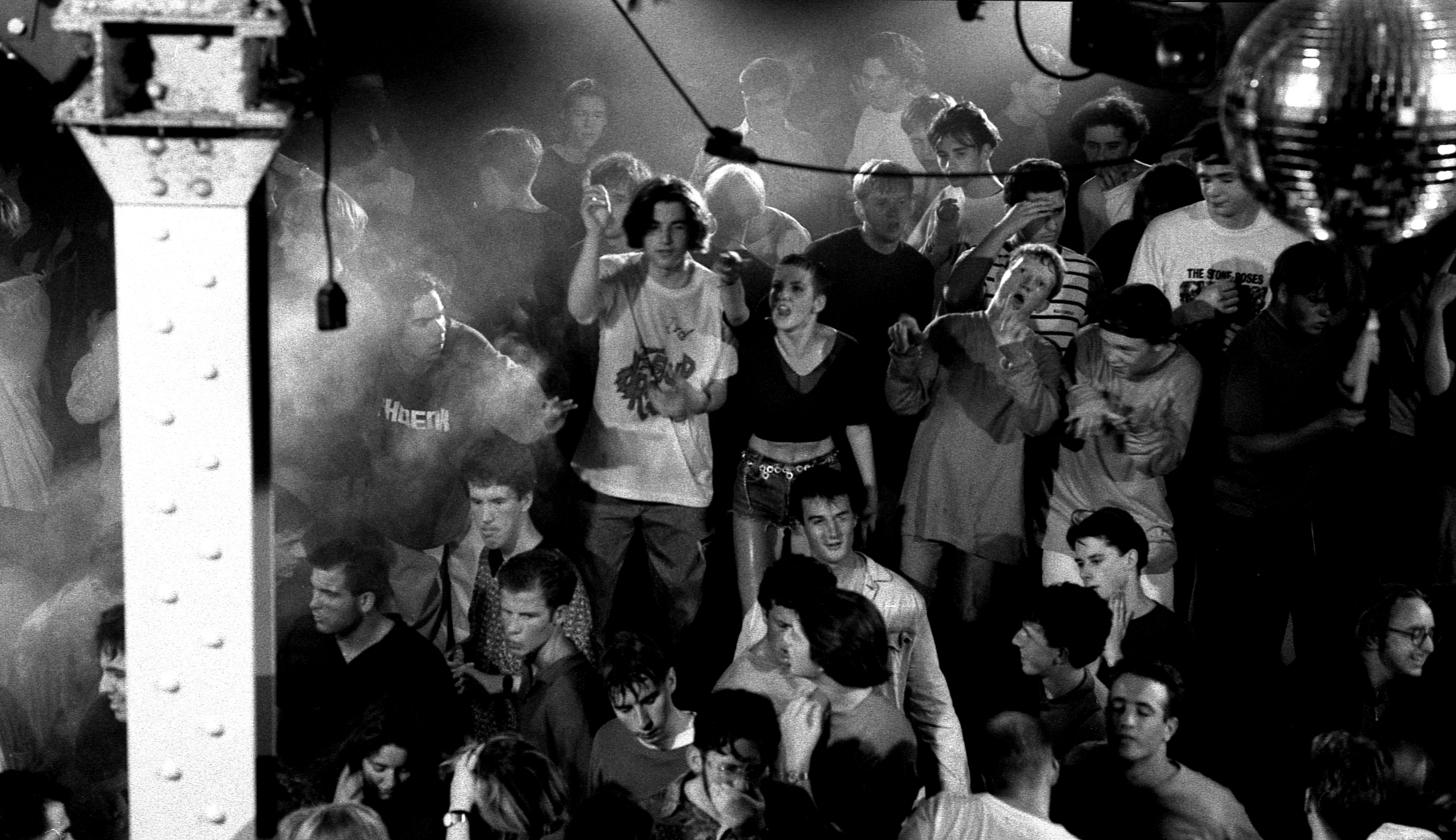 Black and white image of crowded nightclub with people dancing, smoke effects, and stage lighting equipment visible overhead.