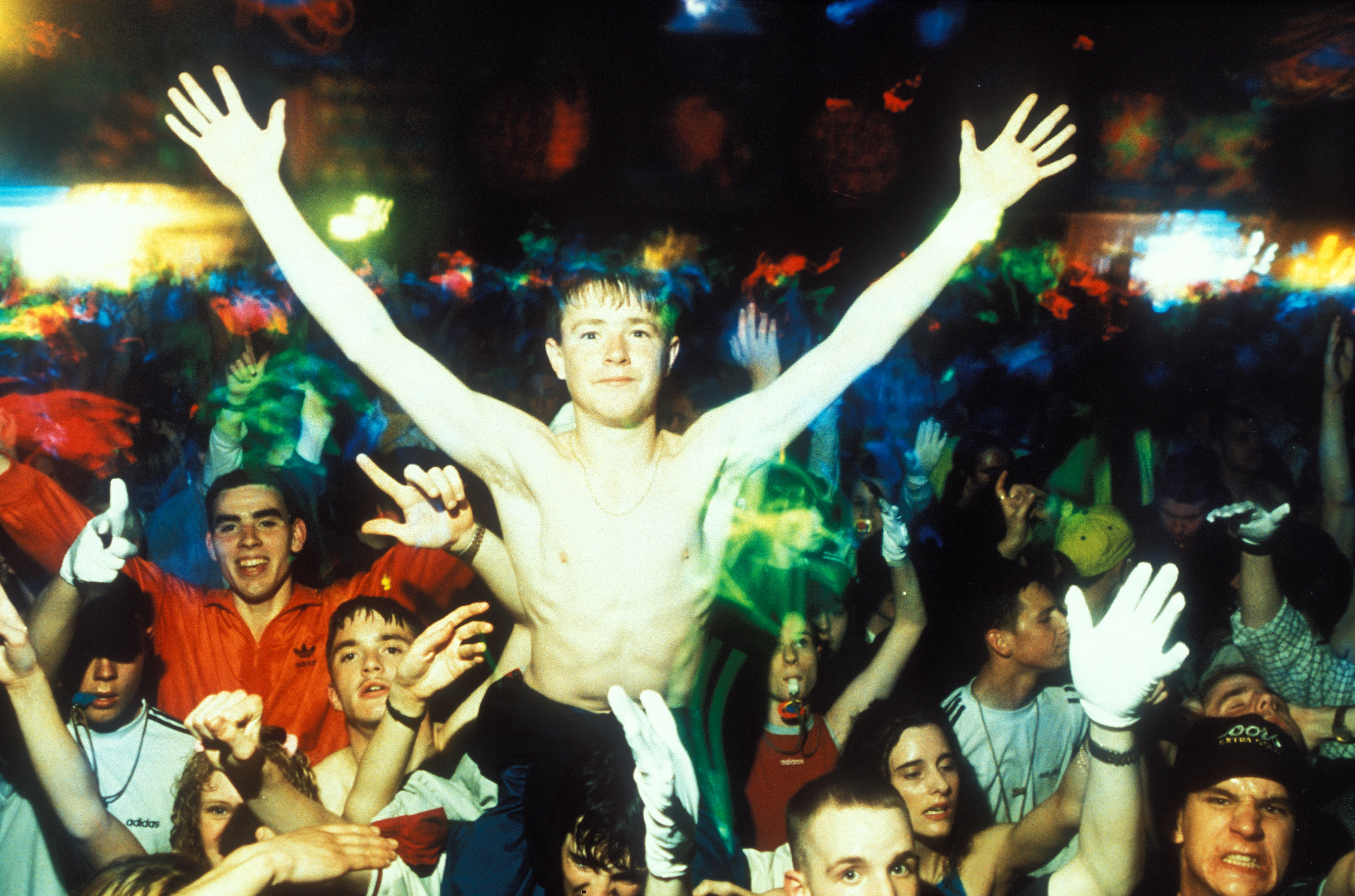 Shirtless man with arms raised high above crowded dancefloor, surrounded by people with hands up, colourful lighting overhead.