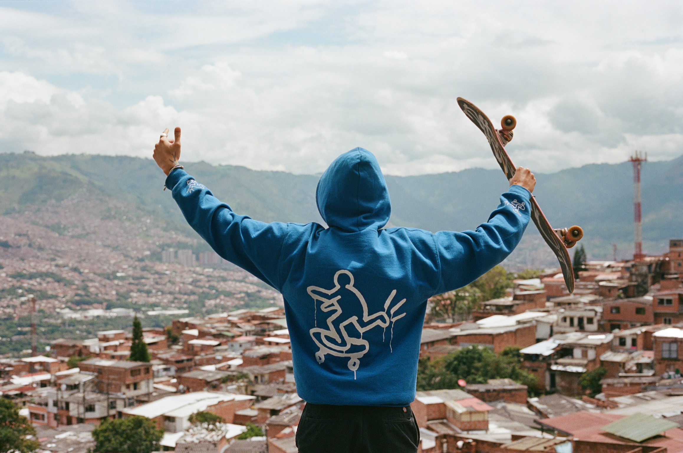 Person in blue hoodie with raised arms holding skateboard, overlooking dense urban cityscape with mountains and cloudy sky.