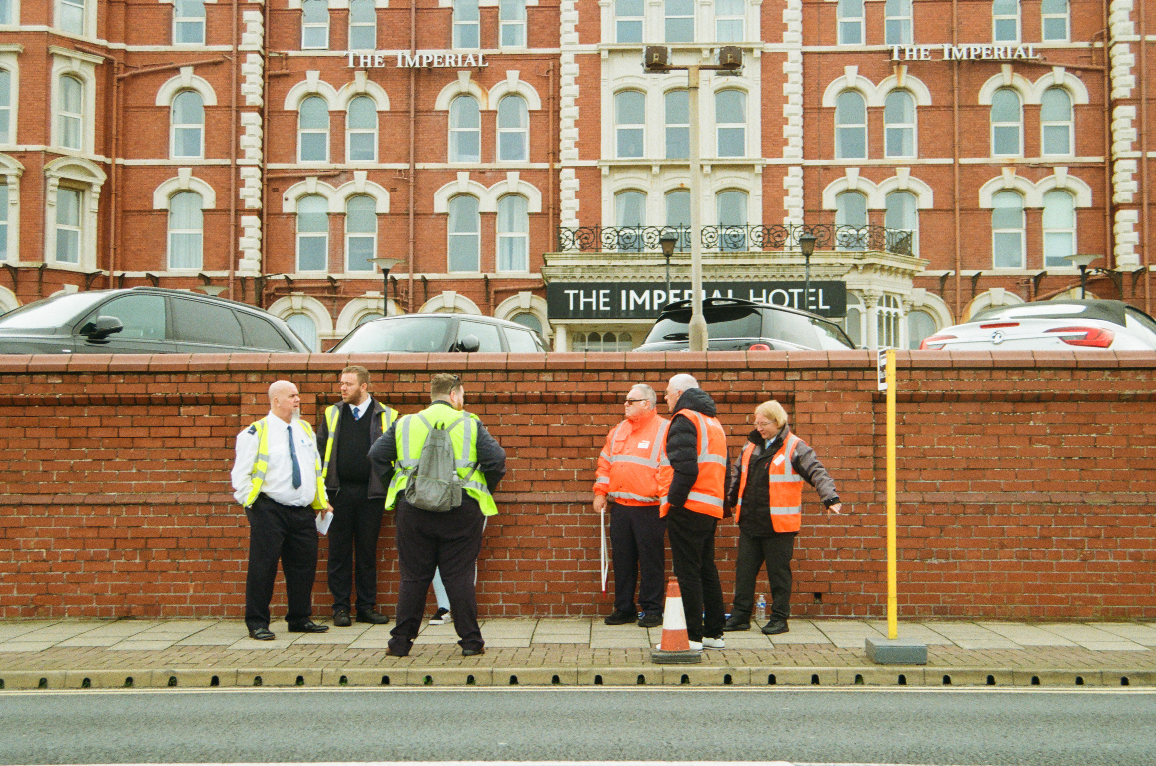 Construction workers in hi-vis jackets stand beside brick wall with parked cars, red brick Imperial Hotel visible in background.