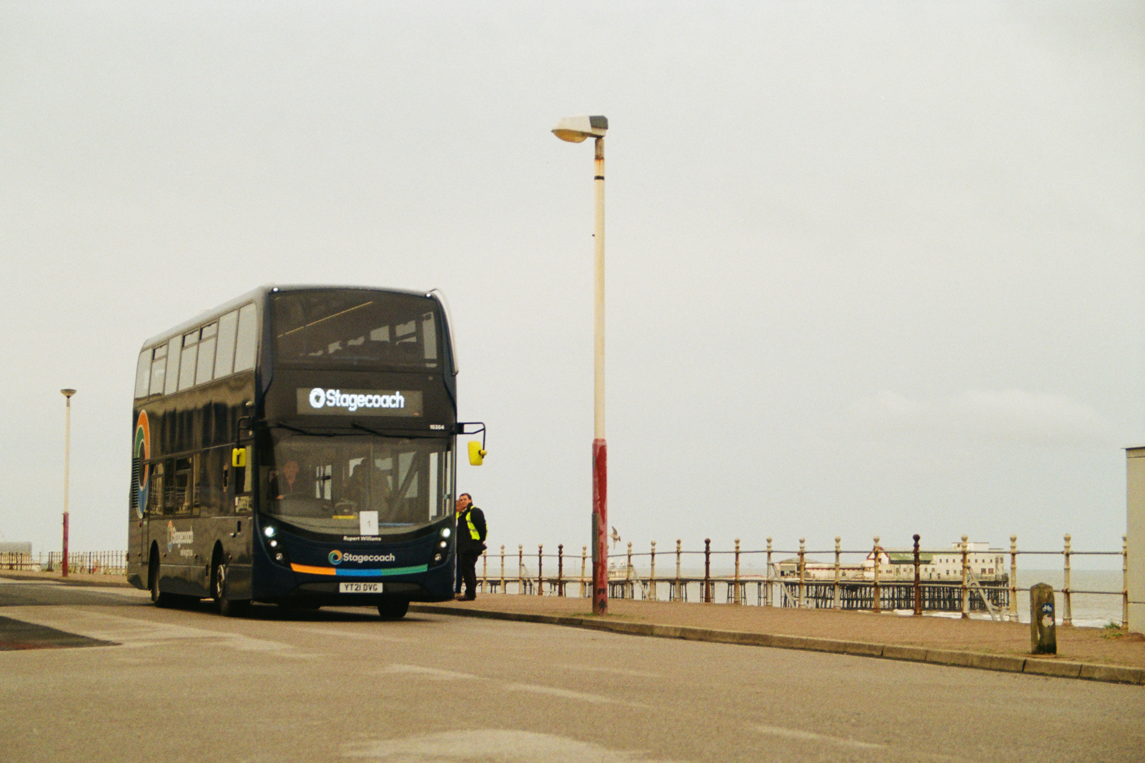 Dark blue double-decker bus displaying "Stagecoach" branding parked on seafront road beside white railings and lamp post under overcast sky.