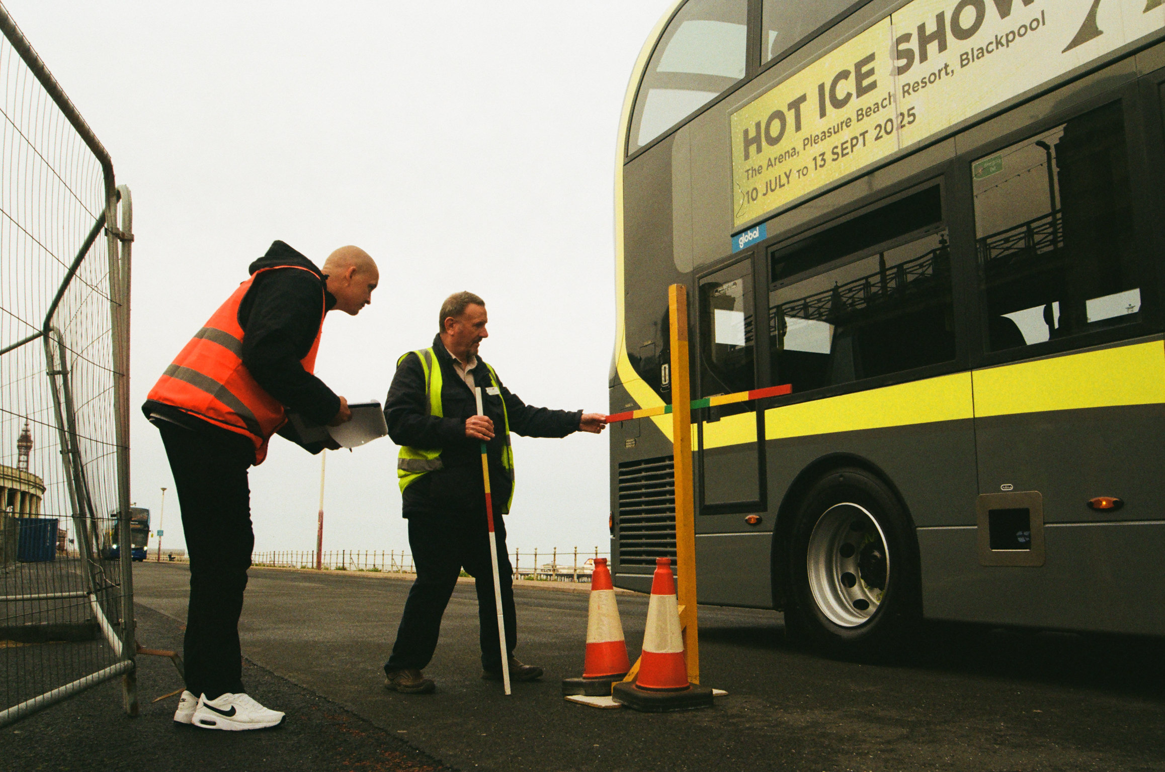 Two workers in high-vis jackets examining yellow bus exterior, traffic cones visible, industrial setting with fence and buildings.