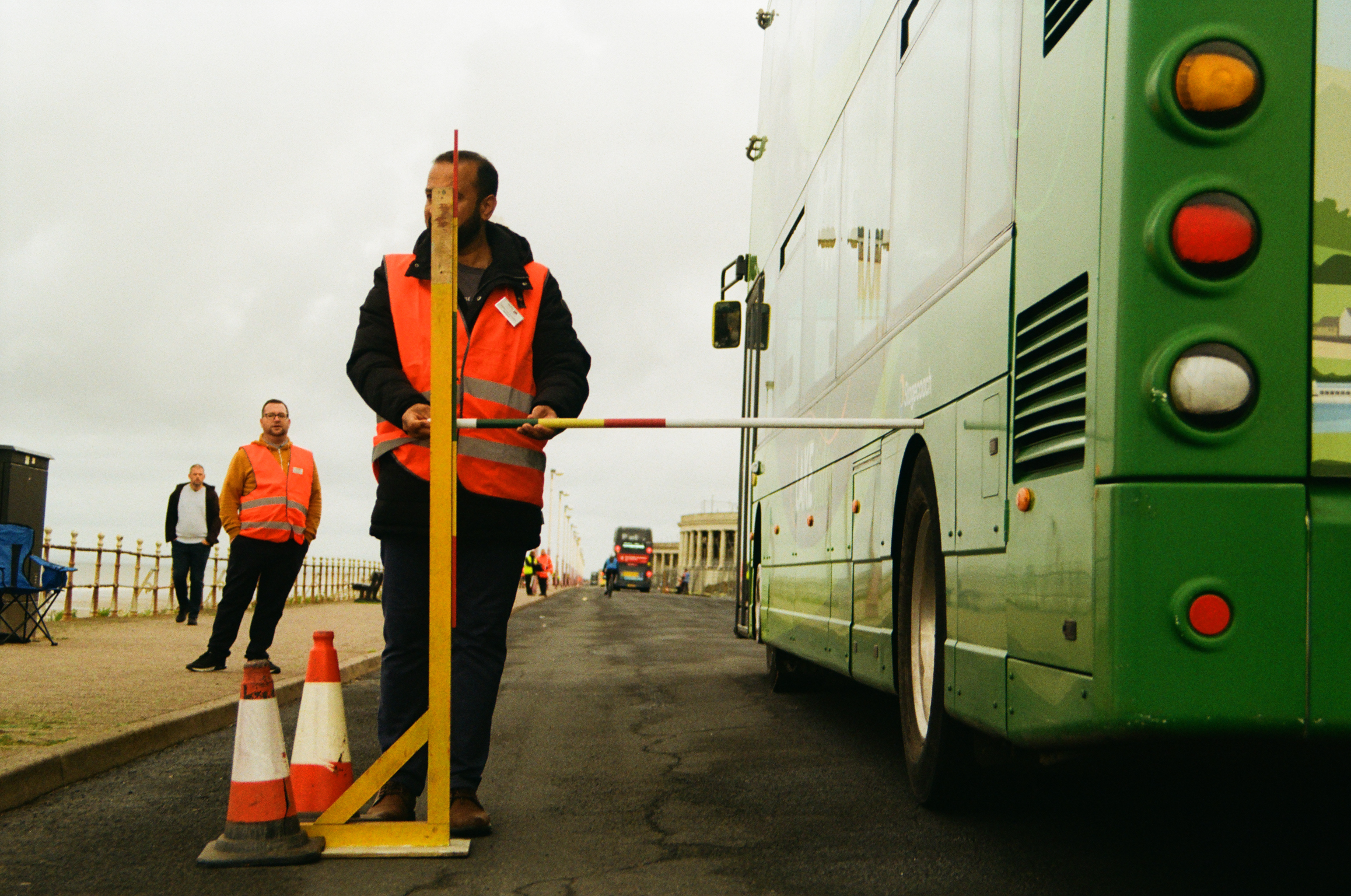 Worker in hi-vis jacket operates yellow pole barrier next to green and white bus. Orange traffic cones and second worker visible in background.