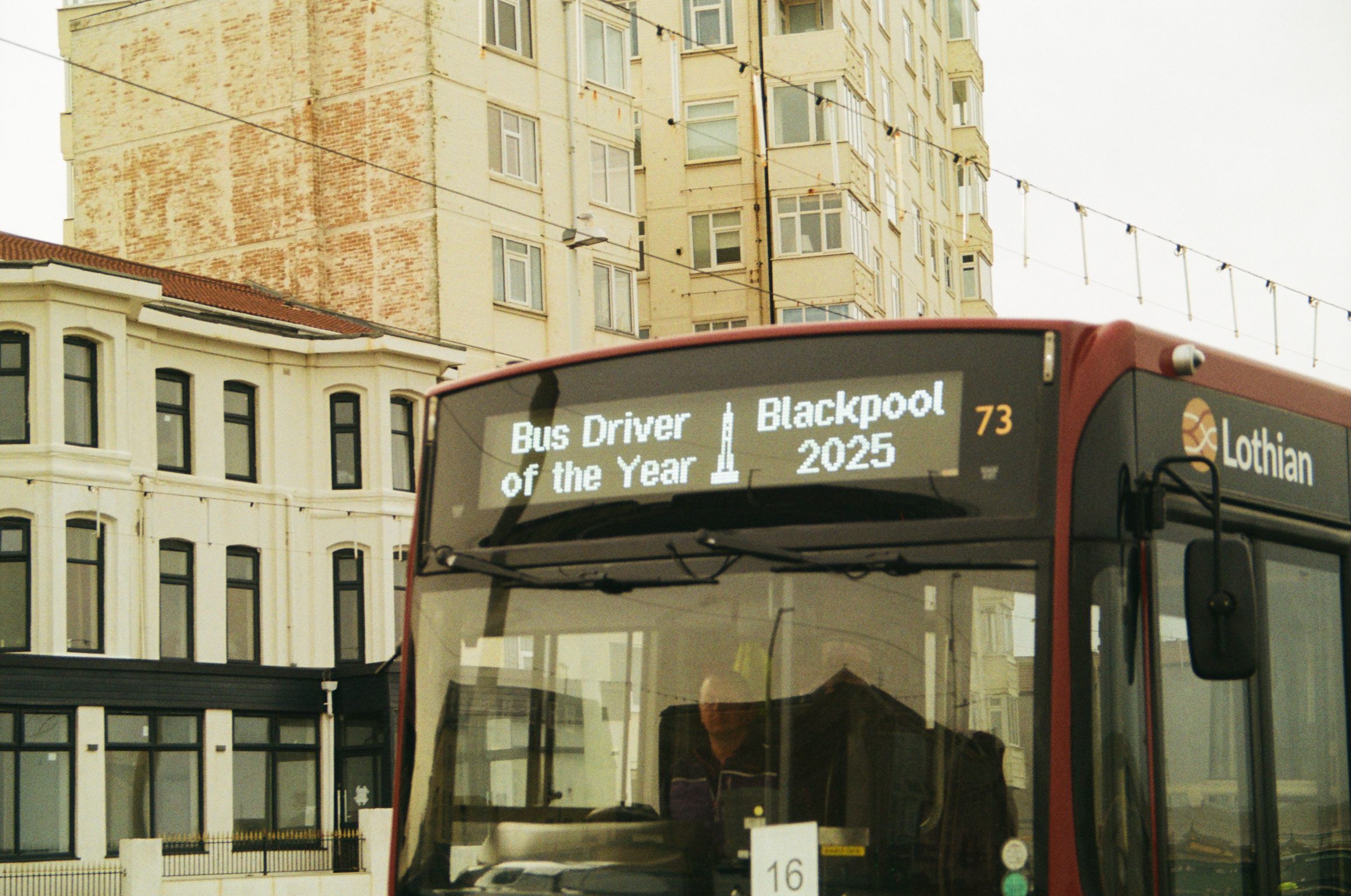 Red Lothian bus displaying "Bus Driver of the Year" and "Blackpool 73 2025" on destination board, parked beside cream-coloured buildings.