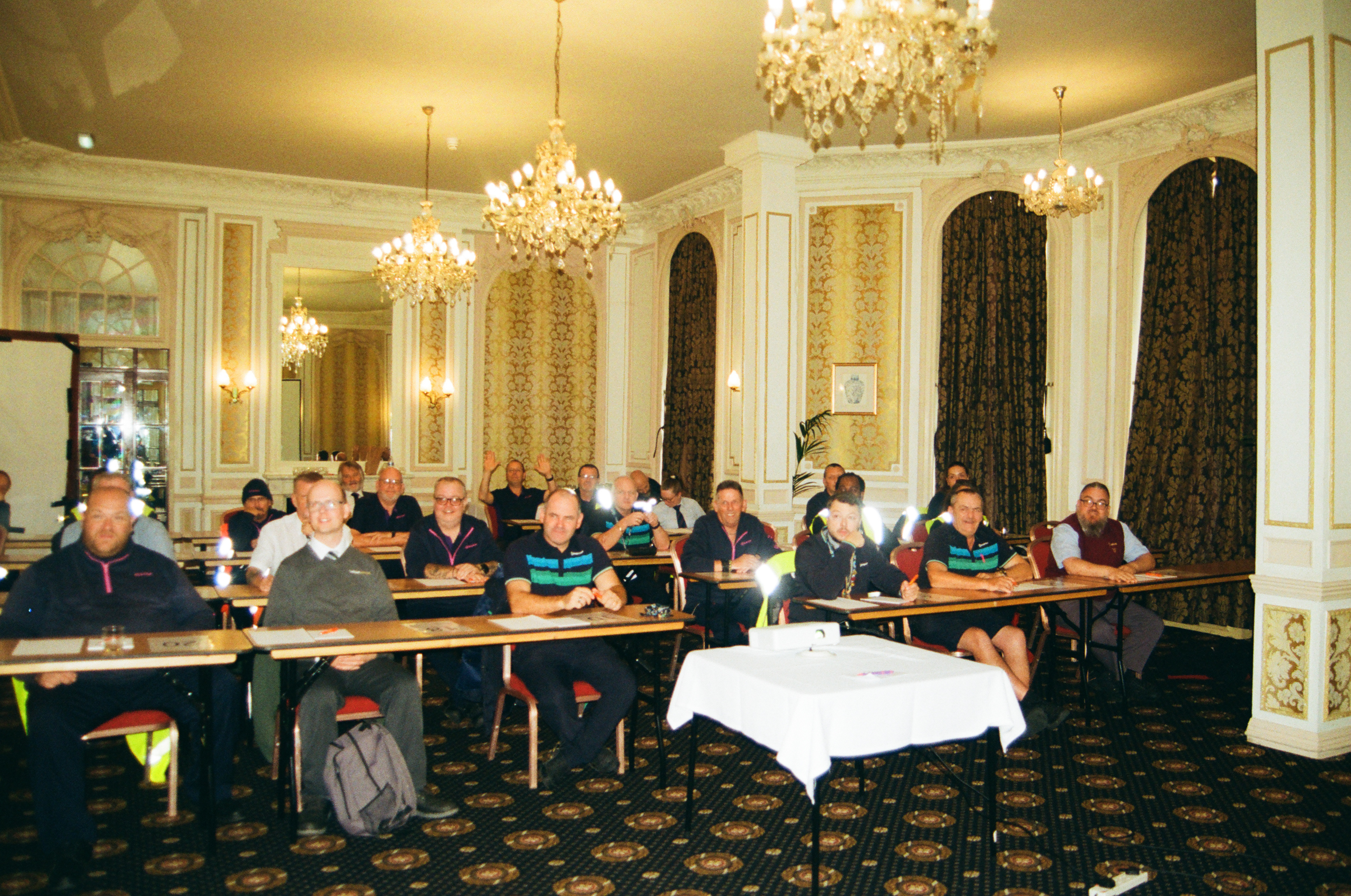 Formal meeting room with ornate chandeliers and gold decor. Multiple attendees seated at wooden tables facing forward.