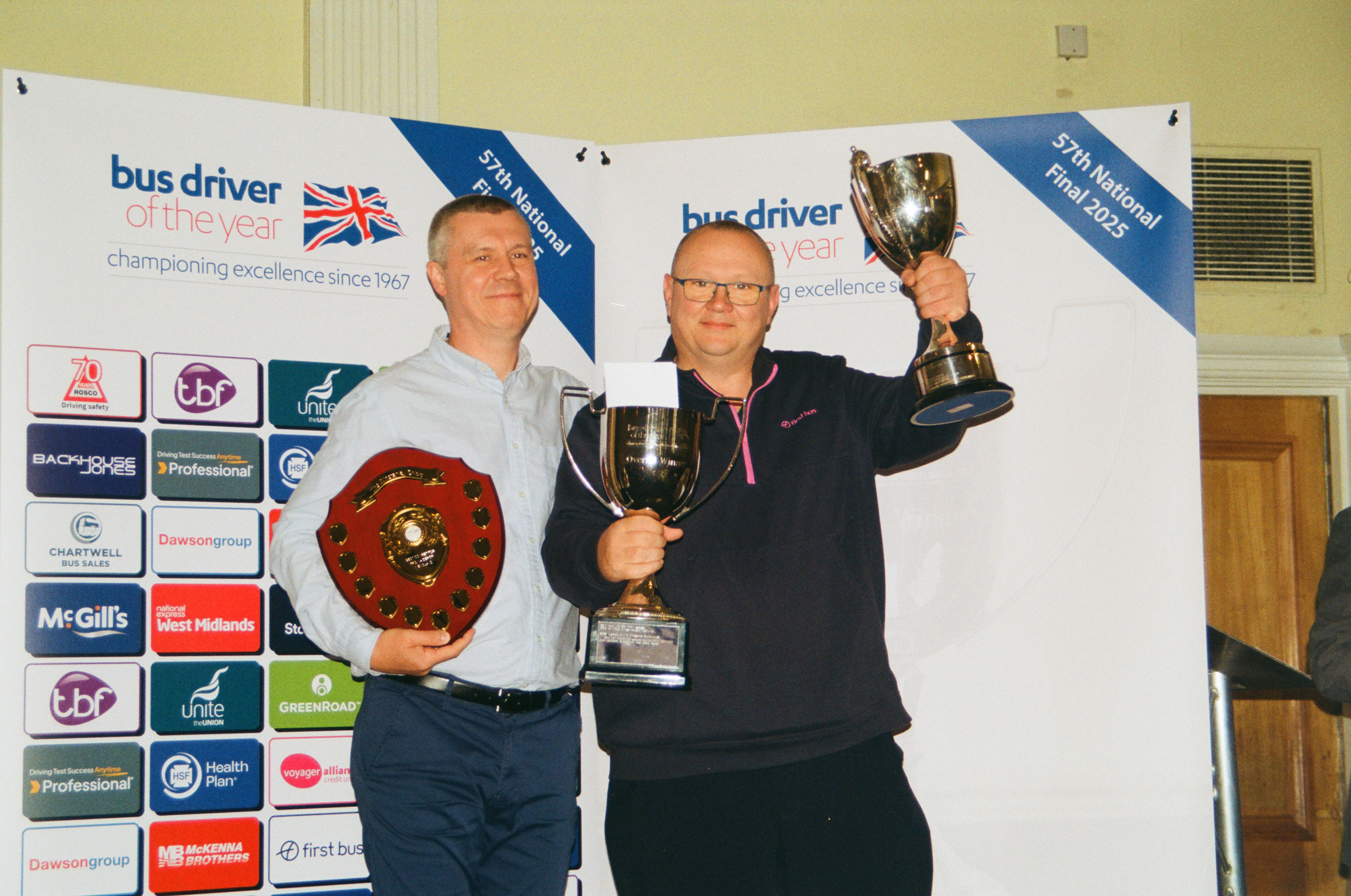 Two men holding trophies at bus driver championship event, standing before white banner with blue sections and sponsor logos.