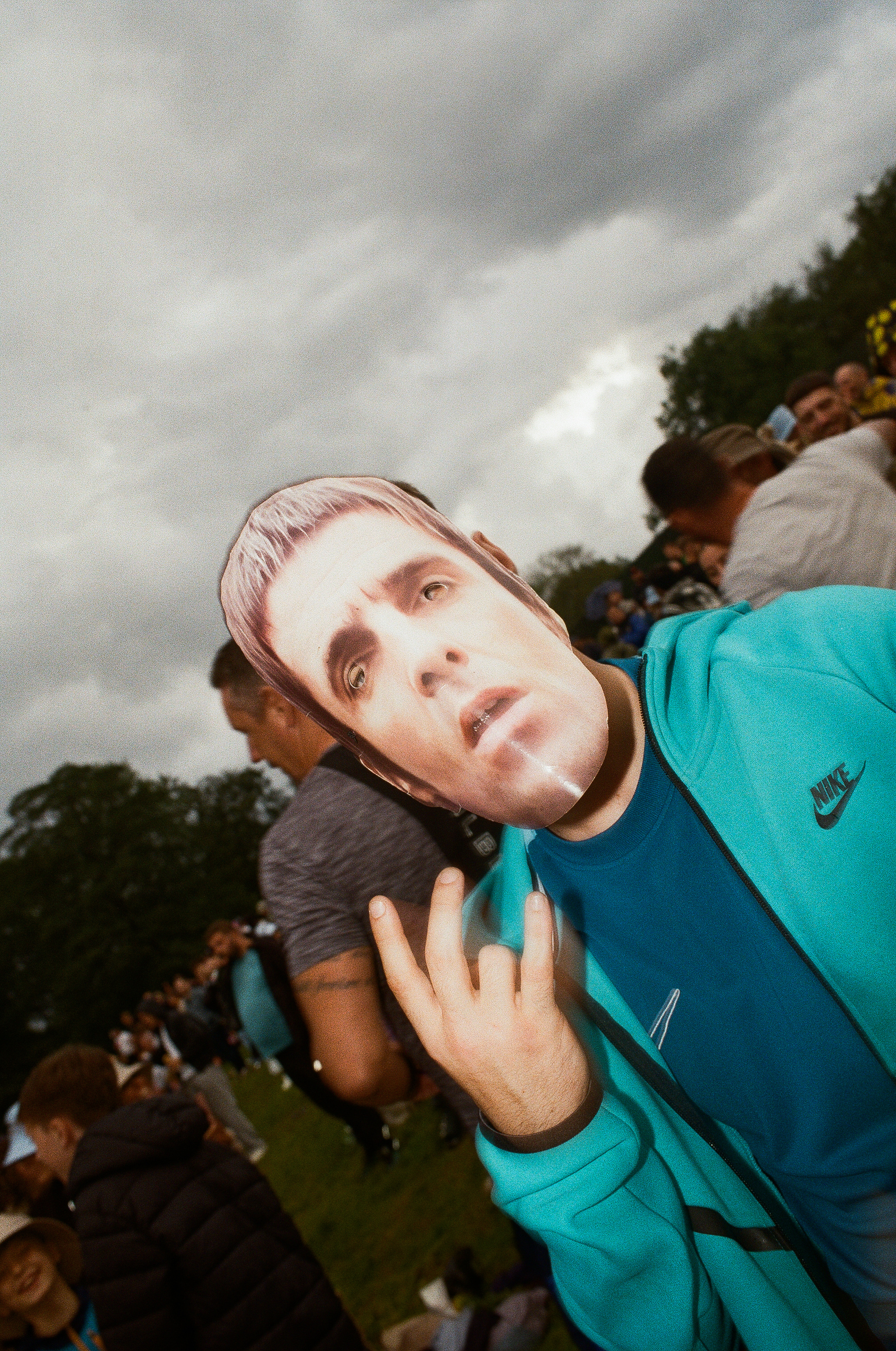 Person in turquoise Nike tracksuit wearing realistic face mask making peace sign gesture, crowd and trees in background under grey sky.