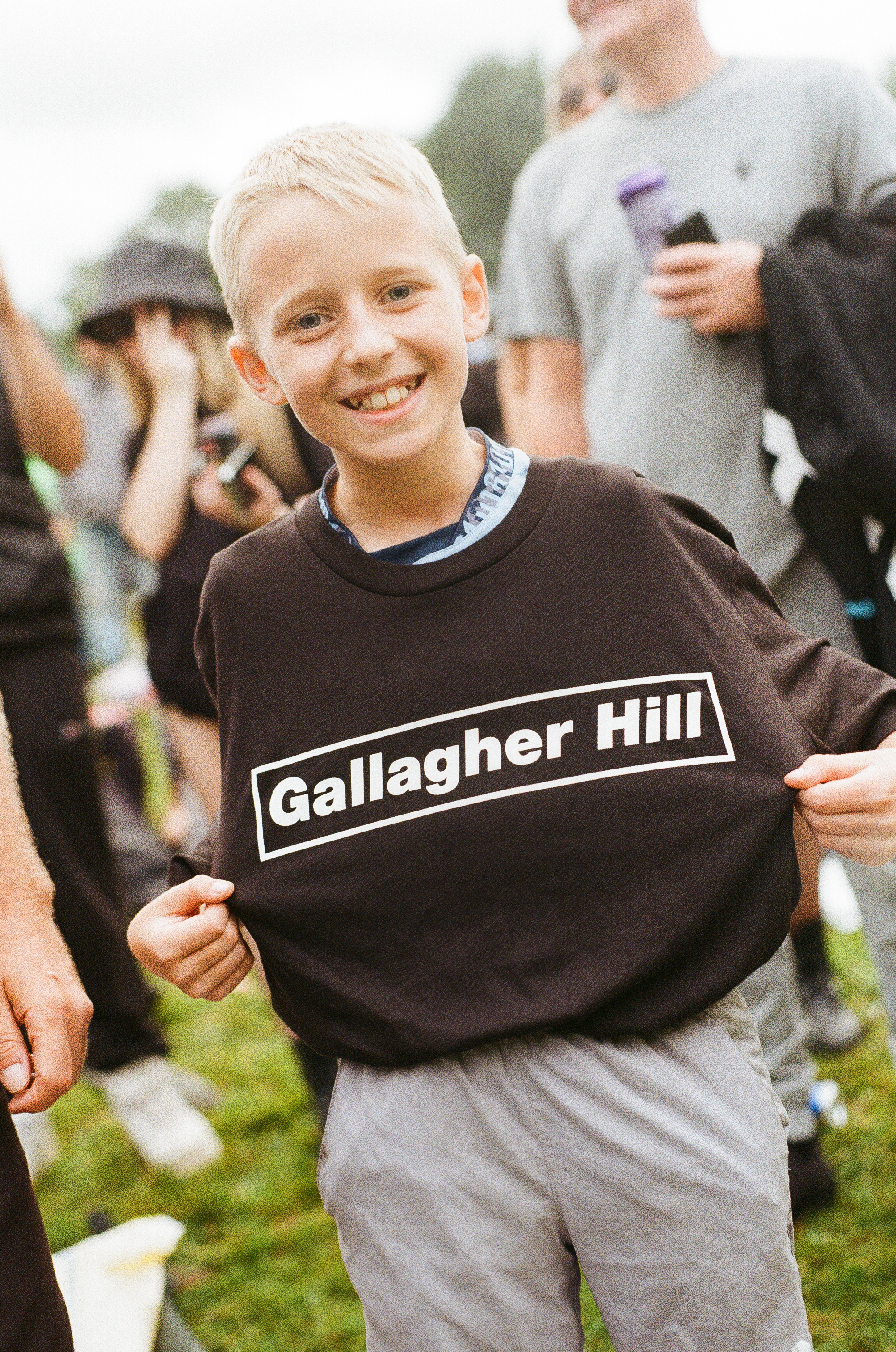 Blonde boy in black Gallagher Hill jumper smiling at camera, with blurred people in background outdoors.