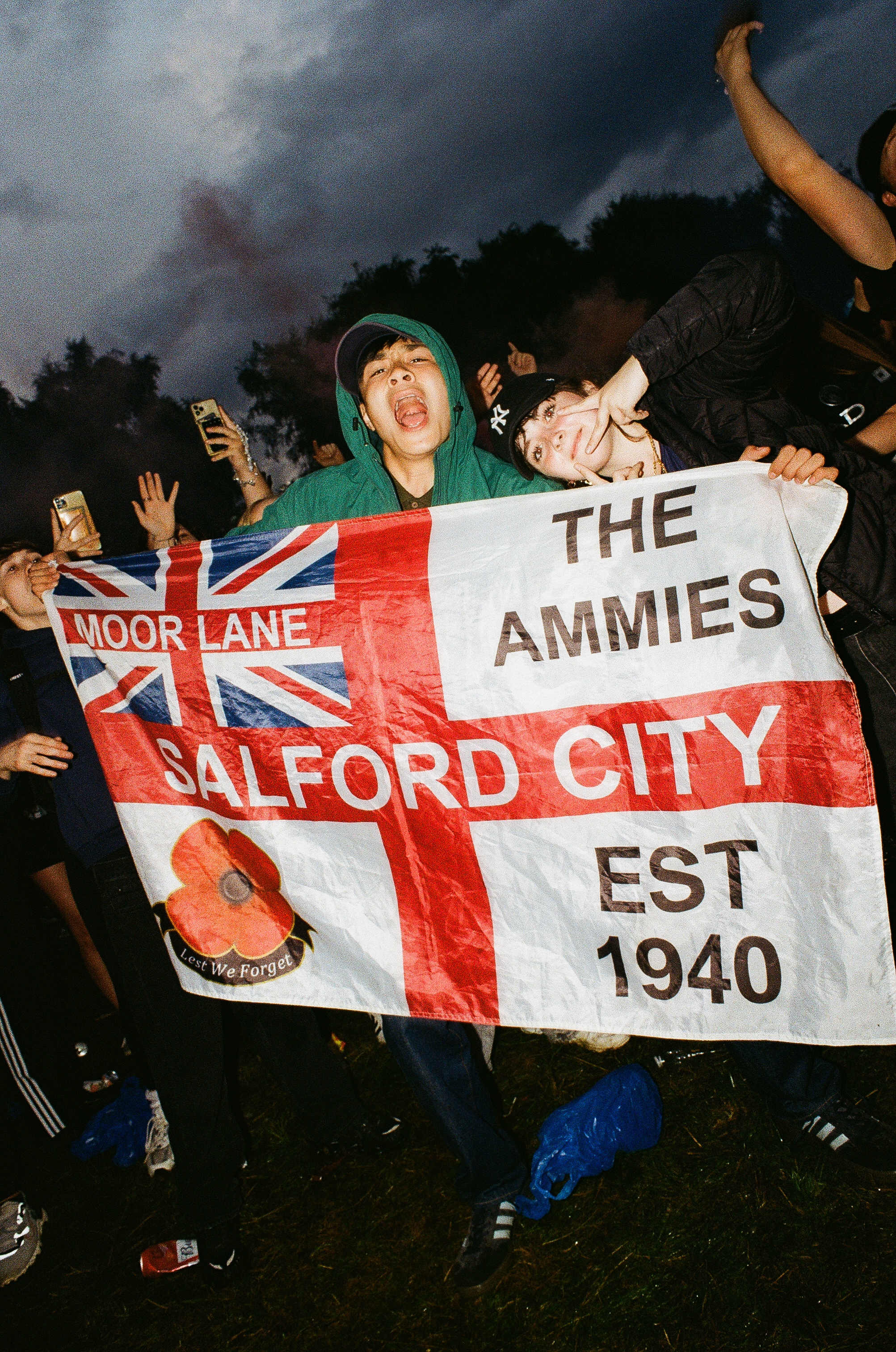 Football supporters holding banner reading "THE AMMIES SALFORD CITY EST 1940" with Union Jack design under stormy evening sky.