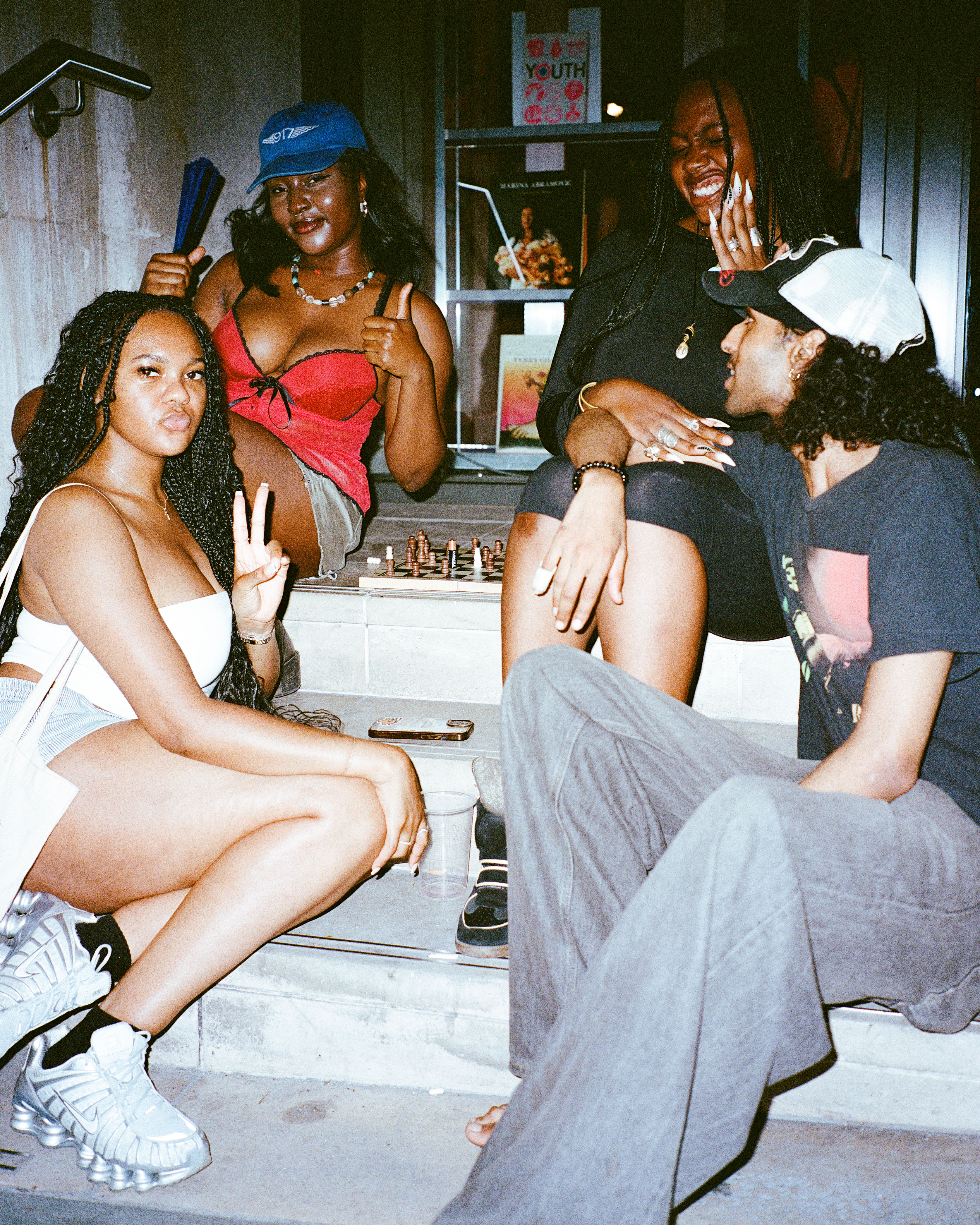 Group of five young women sitting together in casual conversation, some making peace signs, in what appears to be a backstage or dressing room area.